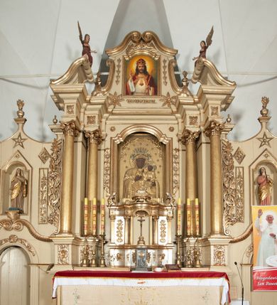 Zdjęcie nr 2: Ołtarz architektoniczny, jednokondygnacyjny, trójosiowy, ze zwieńczeniem. Ołtarz prostopadłościenny, z drewnianą mensą. Antepedium w formie haftowanej tkaniny zdobionej po środku motywem zwieńczonego krzyżem monogramu maryjnego z koroną, ujętego dwunastoma gwiazdami, natomiast w dolnych narożach gałązkami z kwiatami białych lilii.  Tabernakulum z tronem wystawienia w formie jednokondygnacyjnej, trójosiowej bryły architektonicznej, osadzonej na wysokim cokole. Tabernakulum wbudowane w osi środkowej cokołu, drzwiczki prostokątne, zdobione płaskorzeźbionym motywem kielicha eucharystycznego. Flankowane po skrajnych bokach parą pilastrów osie boczne całej struktury są wklęsłe i cofnięte względem osi środkowej ujętej parą kolumienek. Podpory wspierają wyłamujące się ponad nimi belkowanie z gzymsem po środku w formie łuku koszowego. Część środkowa w formie niszy zamkniętej motywem konchy. W zwieńczeniu figurka baranka paschalnego ujętego promienistą glorią, ukazanego na owalnej podstawie osadzonej na 4 wolutowych kabłąkach. Po bokach para figur adorujących aniołów. Cokół zdobiony motywami uskrzydlonych główek anielskich, rocaille’i oraz gałązek z kiściami winnej latorośli. Pola osi bocznych wypełnia para podwieszeń w formie gałązek różanych, partie fryzu pojedyncze rozetki. Struktura tabernakulum i tronu wystawienia jest złocona i srebrzona.   Retabulum osadzone na zdwojonym, zryzalitowanym uskokowo po bokach cokole. Część środkowa ujęta dwoma parami kolumn – wewnętrzne ustawione na tle pary pilastrów, zewnętrzne na tle pary filarów. Podpory dźwigają wyłamujące się ponad nimi belkowanie przerwane w partii architrawu i fryzu. Zwieńczenie ujęte parą spływów wolutowych, zamknięte parą stykających się wolut. Po bokach przerwany, wolutowy przyczółek. W dolnych polach osi bocznych retabulum para półkoliście zamkniętych bramek, ujętych prostą arkadą. Pola osi oddzielone parami stykających się wolut zwieńczonych konchą. W górnej strefie para płytkich niszy zamkniętych półkolistymi łukami nadwieszonymi i dodatkowo ujęta prostym, trójkątnie zamkniętym obramieniem. Osie zwieńczone trzema fragmentami gzymsu oddzielonymi parami wklęsłości.  Pola cokołu osi środkowej retabulum wypełnione ornamentyką roślinną. Część środkowa ujęta parą ażurowych, ornamentalnych  uszaków. Po bokach pola para zwisów w postaci gałązek różanych, w górnych narożach para rozetek. Pola fryzu zdobione rozetami. Pole zwieńczenia w górnej partii dekorowane parą ulistnionych gałązek, po bokach parą kampanuli, dołem w narożach para dużych rozet, pośrodku wić roślinna. Na spływach wolutowych para płomienistych wazonów, a na przyczółku para figur aniołów. Nadłucza bramek osi bocznych zdobione ornamentem roślinnym, nisze ujęte parą stylizowanych kandelabrów, wyżej para rombów z rozetami, pośrodku gwiazda. Na gzymsach wieńczących płomieniste wazony. W polu środkowym obraz w kształcie stojącego prostokąta zamkniętego odcinkowym łukiem nadwieszonym z uskokami, z wizerunkiem Madonny z Dzieciątkiem. W zwieńczeniu obraz w kształcie stojącego prostokąta zamkniętego odcinkowym łukiem nadwieszonym z uskokami, z wizerunkiem Chrystusa Króla. Nisze osi bocznych wypełniają figury, po prawej stronie św. Barbary, po lewej nieznanej świętej. Struktura w kolorze beżowym, detal architektoniczny i ornamentyka złocone.     