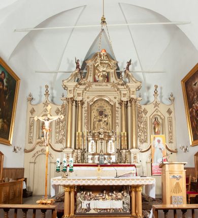 Zdjęcie nr 17: Ołtarz architektoniczny, jednokondygnacyjny, trójosiowy, ze zwieńczeniem. Ołtarz prostopadłościenny, z drewnianą mensą. Antepedium w formie haftowanej tkaniny zdobionej po środku motywem zwieńczonego krzyżem monogramu maryjnego z koroną, ujętego dwunastoma gwiazdami, natomiast w dolnych narożach gałązkami z kwiatami białych lilii.  Tabernakulum z tronem wystawienia w formie jednokondygnacyjnej, trójosiowej bryły architektonicznej, osadzonej na wysokim cokole. Tabernakulum wbudowane w osi środkowej cokołu, drzwiczki prostokątne, zdobione płaskorzeźbionym motywem kielicha eucharystycznego. Flankowane po skrajnych bokach parą pilastrów osie boczne całej struktury są wklęsłe i cofnięte względem osi środkowej ujętej parą kolumienek. Podpory wspierają wyłamujące się ponad nimi belkowanie z gzymsem po środku w formie łuku koszowego. Część środkowa w formie niszy zamkniętej motywem konchy. W zwieńczeniu figurka baranka paschalnego ujętego promienistą glorią, ukazanego na owalnej podstawie osadzonej na 4 wolutowych kabłąkach. Po bokach para figur adorujących aniołów. Cokół zdobiony motywami uskrzydlonych główek anielskich, rocaille’i oraz gałązek z kiściami winnej latorośli. Pola osi bocznych wypełnia para podwieszeń w formie gałązek różanych, partie fryzu pojedyncze rozetki. Struktura tabernakulum i tronu wystawienia jest złocona i srebrzona.   Retabulum osadzone na zdwojonym, zryzalitowanym uskokowo po bokach cokole. Część środkowa ujęta dwoma parami kolumn – wewnętrzne ustawione na tle pary pilastrów, zewnętrzne na tle pary filarów. Podpory dźwigają wyłamujące się ponad nimi belkowanie przerwane w partii architrawu i fryzu. Zwieńczenie ujęte parą spływów wolutowych, zamknięte parą stykających się wolut. Po bokach przerwany, wolutowy przyczółek. W dolnych polach osi bocznych retabulum para półkoliście zamkniętych bramek, ujętych prostą arkadą. Pola osi oddzielone parami stykających się wolut zwieńczonych konchą. W górnej strefie para płytkich niszy zamkniętych półkolistymi łukami nadwieszonymi i dodatkowo ujęta prostym, trójkątnie zamkniętym obramieniem. Osie zwieńczone trzema fragmentami gzymsu oddzielonymi parami wklęsłości.  Pola cokołu osi środkowej retabulum wypełnione ornamentyką roślinną. Część środkowa ujęta parą ażurowych, ornamentalnych  uszaków. Po bokach pola para zwisów w postaci gałązek różanych, w górnych narożach para rozetek. Pola fryzu zdobione rozetami. Pole zwieńczenia w górnej partii dekorowane parą ulistnionych gałązek, po bokach parą kampanuli, dołem w narożach para dużych rozet, pośrodku wić roślinna. Na spływach wolutowych para płomienistych wazonów, a na przyczółku para figur aniołów. Nadłucza bramek osi bocznych zdobione ornamentem roślinnym, nisze ujęte parą stylizowanych kandelabrów, wyżej para rombów z rozetami, pośrodku gwiazda. Na gzymsach wieńczących płomieniste wazony. W polu środkowym obraz w kształcie stojącego prostokąta zamkniętego odcinkowym łukiem nadwieszonym z uskokami, z wizerunkiem Madonny z Dzieciątkiem. W zwieńczeniu obraz w kształcie stojącego prostokąta zamkniętego odcinkowym łukiem nadwieszonym z uskokami, z wizerunkiem Chrystusa Króla. Nisze osi bocznych wypełniają figury, po prawej stronie św. Barbary, po lewej nieznanej świętej. Struktura w kolorze beżowym, detal architektoniczny i ornamentyka złocone.     