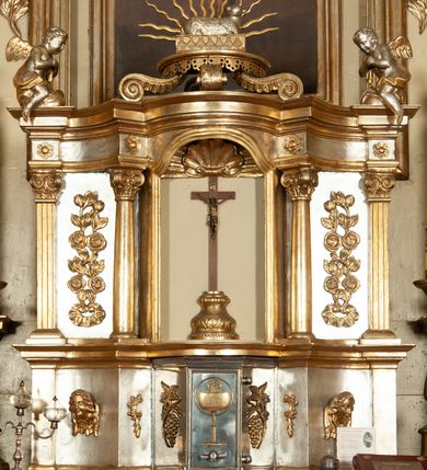 Zdjęcie nr 3: Ołtarz architektoniczny, jednokondygnacyjny, trójosiowy, ze zwieńczeniem. Ołtarz prostopadłościenny, z drewnianą mensą. Antepedium w formie haftowanej tkaniny zdobionej po środku motywem zwieńczonego krzyżem monogramu maryjnego z koroną, ujętego dwunastoma gwiazdami, natomiast w dolnych narożach gałązkami z kwiatami białych lilii.  Tabernakulum z tronem wystawienia w formie jednokondygnacyjnej, trójosiowej bryły architektonicznej, osadzonej na wysokim cokole. Tabernakulum wbudowane w osi środkowej cokołu, drzwiczki prostokątne, zdobione płaskorzeźbionym motywem kielicha eucharystycznego. Flankowane po skrajnych bokach parą pilastrów osie boczne całej struktury są wklęsłe i cofnięte względem osi środkowej ujętej parą kolumienek. Podpory wspierają wyłamujące się ponad nimi belkowanie z gzymsem po środku w formie łuku koszowego. Część środkowa w formie niszy zamkniętej motywem konchy. W zwieńczeniu figurka baranka paschalnego ujętego promienistą glorią, ukazanego na owalnej podstawie osadzonej na 4 wolutowych kabłąkach. Po bokach para figur adorujących aniołów. Cokół zdobiony motywami uskrzydlonych główek anielskich, rocaille’i oraz gałązek z kiściami winnej latorośli. Pola osi bocznych wypełnia para podwieszeń w formie gałązek różanych, partie fryzu pojedyncze rozetki. Struktura tabernakulum i tronu wystawienia jest złocona i srebrzona.   Retabulum osadzone na zdwojonym, zryzalitowanym uskokowo po bokach cokole. Część środkowa ujęta dwoma parami kolumn – wewnętrzne ustawione na tle pary pilastrów, zewnętrzne na tle pary filarów. Podpory dźwigają wyłamujące się ponad nimi belkowanie przerwane w partii architrawu i fryzu. Zwieńczenie ujęte parą spływów wolutowych, zamknięte parą stykających się wolut. Po bokach przerwany, wolutowy przyczółek. W dolnych polach osi bocznych retabulum para półkoliście zamkniętych bramek, ujętych prostą arkadą. Pola osi oddzielone parami stykających się wolut zwieńczonych konchą. W górnej strefie para płytkich niszy zamkniętych półkolistymi łukami nadwieszonymi i dodatkowo ujęta prostym, trójkątnie zamkniętym obramieniem. Osie zwieńczone trzema fragmentami gzymsu oddzielonymi parami wklęsłości.  Pola cokołu osi środkowej retabulum wypełnione ornamentyką roślinną. Część środkowa ujęta parą ażurowych, ornamentalnych  uszaków. Po bokach pola para zwisów w postaci gałązek różanych, w górnych narożach para rozetek. Pola fryzu zdobione rozetami. Pole zwieńczenia w górnej partii dekorowane parą ulistnionych gałązek, po bokach parą kampanuli, dołem w narożach para dużych rozet, pośrodku wić roślinna. Na spływach wolutowych para płomienistych wazonów, a na przyczółku para figur aniołów. Nadłucza bramek osi bocznych zdobione ornamentem roślinnym, nisze ujęte parą stylizowanych kandelabrów, wyżej para rombów z rozetami, pośrodku gwiazda. Na gzymsach wieńczących płomieniste wazony. W polu środkowym obraz w kształcie stojącego prostokąta zamkniętego odcinkowym łukiem nadwieszonym z uskokami, z wizerunkiem Madonny z Dzieciątkiem. W zwieńczeniu obraz w kształcie stojącego prostokąta zamkniętego odcinkowym łukiem nadwieszonym z uskokami, z wizerunkiem Chrystusa Króla. Nisze osi bocznych wypełniają figury, po prawej stronie św. Barbary, po lewej nieznanej świętej. Struktura w kolorze beżowym, detal architektoniczny i ornamentyka złocone.     