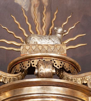 Zdjęcie nr 8: Ołtarz architektoniczny, jednokondygnacyjny, trójosiowy, ze zwieńczeniem. Ołtarz prostopadłościenny, z drewnianą mensą. Antepedium w formie haftowanej tkaniny zdobionej po środku motywem zwieńczonego krzyżem monogramu maryjnego z koroną, ujętego dwunastoma gwiazdami, natomiast w dolnych narożach gałązkami z kwiatami białych lilii.  Tabernakulum z tronem wystawienia w formie jednokondygnacyjnej, trójosiowej bryły architektonicznej, osadzonej na wysokim cokole. Tabernakulum wbudowane w osi środkowej cokołu, drzwiczki prostokątne, zdobione płaskorzeźbionym motywem kielicha eucharystycznego. Flankowane po skrajnych bokach parą pilastrów osie boczne całej struktury są wklęsłe i cofnięte względem osi środkowej ujętej parą kolumienek. Podpory wspierają wyłamujące się ponad nimi belkowanie z gzymsem po środku w formie łuku koszowego. Część środkowa w formie niszy zamkniętej motywem konchy. W zwieńczeniu figurka baranka paschalnego ujętego promienistą glorią, ukazanego na owalnej podstawie osadzonej na 4 wolutowych kabłąkach. Po bokach para figur adorujących aniołów. Cokół zdobiony motywami uskrzydlonych główek anielskich, rocaille’i oraz gałązek z kiściami winnej latorośli. Pola osi bocznych wypełnia para podwieszeń w formie gałązek różanych, partie fryzu pojedyncze rozetki. Struktura tabernakulum i tronu wystawienia jest złocona i srebrzona.   Retabulum osadzone na zdwojonym, zryzalitowanym uskokowo po bokach cokole. Część środkowa ujęta dwoma parami kolumn – wewnętrzne ustawione na tle pary pilastrów, zewnętrzne na tle pary filarów. Podpory dźwigają wyłamujące się ponad nimi belkowanie przerwane w partii architrawu i fryzu. Zwieńczenie ujęte parą spływów wolutowych, zamknięte parą stykających się wolut. Po bokach przerwany, wolutowy przyczółek. W dolnych polach osi bocznych retabulum para półkoliście zamkniętych bramek, ujętych prostą arkadą. Pola osi oddzielone parami stykających się wolut zwieńczonych konchą. W górnej strefie para płytkich niszy zamkniętych półkolistymi łukami nadwieszonymi i dodatkowo ujęta prostym, trójkątnie zamkniętym obramieniem. Osie zwieńczone trzema fragmentami gzymsu oddzielonymi parami wklęsłości.  Pola cokołu osi środkowej retabulum wypełnione ornamentyką roślinną. Część środkowa ujęta parą ażurowych, ornamentalnych  uszaków. Po bokach pola para zwisów w postaci gałązek różanych, w górnych narożach para rozetek. Pola fryzu zdobione rozetami. Pole zwieńczenia w górnej partii dekorowane parą ulistnionych gałązek, po bokach parą kampanuli, dołem w narożach para dużych rozet, pośrodku wić roślinna. Na spływach wolutowych para płomienistych wazonów, a na przyczółku para figur aniołów. Nadłucza bramek osi bocznych zdobione ornamentem roślinnym, nisze ujęte parą stylizowanych kandelabrów, wyżej para rombów z rozetami, pośrodku gwiazda. Na gzymsach wieńczących płomieniste wazony. W polu środkowym obraz w kształcie stojącego prostokąta zamkniętego odcinkowym łukiem nadwieszonym z uskokami, z wizerunkiem Madonny z Dzieciątkiem. W zwieńczeniu obraz w kształcie stojącego prostokąta zamkniętego odcinkowym łukiem nadwieszonym z uskokami, z wizerunkiem Chrystusa Króla. Nisze osi bocznych wypełniają figury, po prawej stronie św. Barbary, po lewej nieznanej świętej. Struktura w kolorze beżowym, detal architektoniczny i ornamentyka złocone.     