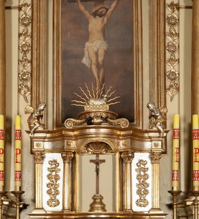 Zdjęcie nr 20: Ołtarz architektoniczny, jednokondygnacyjny, trójosiowy, ze zwieńczeniem. Ołtarz prostopadłościenny, z drewnianą mensą. Antepedium w formie haftowanej tkaniny zdobionej po środku motywem zwieńczonego krzyżem monogramu maryjnego z koroną, ujętego dwunastoma gwiazdami, natomiast w dolnych narożach gałązkami z kwiatami białych lilii.  Tabernakulum z tronem wystawienia w formie jednokondygnacyjnej, trójosiowej bryły architektonicznej, osadzonej na wysokim cokole. Tabernakulum wbudowane w osi środkowej cokołu, drzwiczki prostokątne, zdobione płaskorzeźbionym motywem kielicha eucharystycznego. Flankowane po skrajnych bokach parą pilastrów osie boczne całej struktury są wklęsłe i cofnięte względem osi środkowej ujętej parą kolumienek. Podpory wspierają wyłamujące się ponad nimi belkowanie z gzymsem po środku w formie łuku koszowego. Część środkowa w formie niszy zamkniętej motywem konchy. W zwieńczeniu figurka baranka paschalnego ujętego promienistą glorią, ukazanego na owalnej podstawie osadzonej na 4 wolutowych kabłąkach. Po bokach para figur adorujących aniołów. Cokół zdobiony motywami uskrzydlonych główek anielskich, rocaille’i oraz gałązek z kiściami winnej latorośli. Pola osi bocznych wypełnia para podwieszeń w formie gałązek różanych, partie fryzu pojedyncze rozetki. Struktura tabernakulum i tronu wystawienia jest złocona i srebrzona.   Retabulum osadzone na zdwojonym, zryzalitowanym uskokowo po bokach cokole. Część środkowa ujęta dwoma parami kolumn – wewnętrzne ustawione na tle pary pilastrów, zewnętrzne na tle pary filarów. Podpory dźwigają wyłamujące się ponad nimi belkowanie przerwane w partii architrawu i fryzu. Zwieńczenie ujęte parą spływów wolutowych, zamknięte parą stykających się wolut. Po bokach przerwany, wolutowy przyczółek. W dolnych polach osi bocznych retabulum para półkoliście zamkniętych bramek, ujętych prostą arkadą. Pola osi oddzielone parami stykających się wolut zwieńczonych konchą. W górnej strefie para płytkich niszy zamkniętych półkolistymi łukami nadwieszonymi i dodatkowo ujęta prostym, trójkątnie zamkniętym obramieniem. Osie zwieńczone trzema fragmentami gzymsu oddzielonymi parami wklęsłości.  Pola cokołu osi środkowej retabulum wypełnione ornamentyką roślinną. Część środkowa ujęta parą ażurowych, ornamentalnych  uszaków. Po bokach pola para zwisów w postaci gałązek różanych, w górnych narożach para rozetek. Pola fryzu zdobione rozetami. Pole zwieńczenia w górnej partii dekorowane parą ulistnionych gałązek, po bokach parą kampanuli, dołem w narożach para dużych rozet, pośrodku wić roślinna. Na spływach wolutowych para płomienistych wazonów, a na przyczółku para figur aniołów. Nadłucza bramek osi bocznych zdobione ornamentem roślinnym, nisze ujęte parą stylizowanych kandelabrów, wyżej para rombów z rozetami, pośrodku gwiazda. Na gzymsach wieńczących płomieniste wazony. W polu środkowym obraz w kształcie stojącego prostokąta zamkniętego odcinkowym łukiem nadwieszonym z uskokami, z wizerunkiem Madonny z Dzieciątkiem. W zwieńczeniu obraz w kształcie stojącego prostokąta zamkniętego odcinkowym łukiem nadwieszonym z uskokami, z wizerunkiem Chrystusa Króla. Nisze osi bocznych wypełniają figury, po prawej stronie św. Barbary, po lewej nieznanej świętej. Struktura w kolorze beżowym, detal architektoniczny i ornamentyka złocone.     