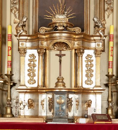 Zdjęcie nr 6: Ołtarz architektoniczny, jednokondygnacyjny, trójosiowy, ze zwieńczeniem. Ołtarz prostopadłościenny, z drewnianą mensą. Antepedium w formie haftowanej tkaniny zdobionej po środku motywem zwieńczonego krzyżem monogramu maryjnego z koroną, ujętego dwunastoma gwiazdami, natomiast w dolnych narożach gałązkami z kwiatami białych lilii.  Tabernakulum z tronem wystawienia w formie jednokondygnacyjnej, trójosiowej bryły architektonicznej, osadzonej na wysokim cokole. Tabernakulum wbudowane w osi środkowej cokołu, drzwiczki prostokątne, zdobione płaskorzeźbionym motywem kielicha eucharystycznego. Flankowane po skrajnych bokach parą pilastrów osie boczne całej struktury są wklęsłe i cofnięte względem osi środkowej ujętej parą kolumienek. Podpory wspierają wyłamujące się ponad nimi belkowanie z gzymsem po środku w formie łuku koszowego. Część środkowa w formie niszy zamkniętej motywem konchy. W zwieńczeniu figurka baranka paschalnego ujętego promienistą glorią, ukazanego na owalnej podstawie osadzonej na 4 wolutowych kabłąkach. Po bokach para figur adorujących aniołów. Cokół zdobiony motywami uskrzydlonych główek anielskich, rocaille’i oraz gałązek z kiściami winnej latorośli. Pola osi bocznych wypełnia para podwieszeń w formie gałązek różanych, partie fryzu pojedyncze rozetki. Struktura tabernakulum i tronu wystawienia jest złocona i srebrzona.   Retabulum osadzone na zdwojonym, zryzalitowanym uskokowo po bokach cokole. Część środkowa ujęta dwoma parami kolumn – wewnętrzne ustawione na tle pary pilastrów, zewnętrzne na tle pary filarów. Podpory dźwigają wyłamujące się ponad nimi belkowanie przerwane w partii architrawu i fryzu. Zwieńczenie ujęte parą spływów wolutowych, zamknięte parą stykających się wolut. Po bokach przerwany, wolutowy przyczółek. W dolnych polach osi bocznych retabulum para półkoliście zamkniętych bramek, ujętych prostą arkadą. Pola osi oddzielone parami stykających się wolut zwieńczonych konchą. W górnej strefie para płytkich niszy zamkniętych półkolistymi łukami nadwieszonymi i dodatkowo ujęta prostym, trójkątnie zamkniętym obramieniem. Osie zwieńczone trzema fragmentami gzymsu oddzielonymi parami wklęsłości.  Pola cokołu osi środkowej retabulum wypełnione ornamentyką roślinną. Część środkowa ujęta parą ażurowych, ornamentalnych  uszaków. Po bokach pola para zwisów w postaci gałązek różanych, w górnych narożach para rozetek. Pola fryzu zdobione rozetami. Pole zwieńczenia w górnej partii dekorowane parą ulistnionych gałązek, po bokach parą kampanuli, dołem w narożach para dużych rozet, pośrodku wić roślinna. Na spływach wolutowych para płomienistych wazonów, a na przyczółku para figur aniołów. Nadłucza bramek osi bocznych zdobione ornamentem roślinnym, nisze ujęte parą stylizowanych kandelabrów, wyżej para rombów z rozetami, pośrodku gwiazda. Na gzymsach wieńczących płomieniste wazony. W polu środkowym obraz w kształcie stojącego prostokąta zamkniętego odcinkowym łukiem nadwieszonym z uskokami, z wizerunkiem Madonny z Dzieciątkiem. W zwieńczeniu obraz w kształcie stojącego prostokąta zamkniętego odcinkowym łukiem nadwieszonym z uskokami, z wizerunkiem Chrystusa Króla. Nisze osi bocznych wypełniają figury, po prawej stronie św. Barbary, po lewej nieznanej świętej. Struktura w kolorze beżowym, detal architektoniczny i ornamentyka złocone.     