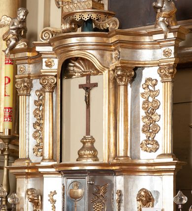 Zdjęcie nr 7: Ołtarz architektoniczny, jednokondygnacyjny, trójosiowy, ze zwieńczeniem. Ołtarz prostopadłościenny, z drewnianą mensą. Antepedium w formie haftowanej tkaniny zdobionej po środku motywem zwieńczonego krzyżem monogramu maryjnego z koroną, ujętego dwunastoma gwiazdami, natomiast w dolnych narożach gałązkami z kwiatami białych lilii.  Tabernakulum z tronem wystawienia w formie jednokondygnacyjnej, trójosiowej bryły architektonicznej, osadzonej na wysokim cokole. Tabernakulum wbudowane w osi środkowej cokołu, drzwiczki prostokątne, zdobione płaskorzeźbionym motywem kielicha eucharystycznego. Flankowane po skrajnych bokach parą pilastrów osie boczne całej struktury są wklęsłe i cofnięte względem osi środkowej ujętej parą kolumienek. Podpory wspierają wyłamujące się ponad nimi belkowanie z gzymsem po środku w formie łuku koszowego. Część środkowa w formie niszy zamkniętej motywem konchy. W zwieńczeniu figurka baranka paschalnego ujętego promienistą glorią, ukazanego na owalnej podstawie osadzonej na 4 wolutowych kabłąkach. Po bokach para figur adorujących aniołów. Cokół zdobiony motywami uskrzydlonych główek anielskich, rocaille’i oraz gałązek z kiściami winnej latorośli. Pola osi bocznych wypełnia para podwieszeń w formie gałązek różanych, partie fryzu pojedyncze rozetki. Struktura tabernakulum i tronu wystawienia jest złocona i srebrzona.   Retabulum osadzone na zdwojonym, zryzalitowanym uskokowo po bokach cokole. Część środkowa ujęta dwoma parami kolumn – wewnętrzne ustawione na tle pary pilastrów, zewnętrzne na tle pary filarów. Podpory dźwigają wyłamujące się ponad nimi belkowanie przerwane w partii architrawu i fryzu. Zwieńczenie ujęte parą spływów wolutowych, zamknięte parą stykających się wolut. Po bokach przerwany, wolutowy przyczółek. W dolnych polach osi bocznych retabulum para półkoliście zamkniętych bramek, ujętych prostą arkadą. Pola osi oddzielone parami stykających się wolut zwieńczonych konchą. W górnej strefie para płytkich niszy zamkniętych półkolistymi łukami nadwieszonymi i dodatkowo ujęta prostym, trójkątnie zamkniętym obramieniem. Osie zwieńczone trzema fragmentami gzymsu oddzielonymi parami wklęsłości.  Pola cokołu osi środkowej retabulum wypełnione ornamentyką roślinną. Część środkowa ujęta parą ażurowych, ornamentalnych  uszaków. Po bokach pola para zwisów w postaci gałązek różanych, w górnych narożach para rozetek. Pola fryzu zdobione rozetami. Pole zwieńczenia w górnej partii dekorowane parą ulistnionych gałązek, po bokach parą kampanuli, dołem w narożach para dużych rozet, pośrodku wić roślinna. Na spływach wolutowych para płomienistych wazonów, a na przyczółku para figur aniołów. Nadłucza bramek osi bocznych zdobione ornamentem roślinnym, nisze ujęte parą stylizowanych kandelabrów, wyżej para rombów z rozetami, pośrodku gwiazda. Na gzymsach wieńczących płomieniste wazony. W polu środkowym obraz w kształcie stojącego prostokąta zamkniętego odcinkowym łukiem nadwieszonym z uskokami, z wizerunkiem Madonny z Dzieciątkiem. W zwieńczeniu obraz w kształcie stojącego prostokąta zamkniętego odcinkowym łukiem nadwieszonym z uskokami, z wizerunkiem Chrystusa Króla. Nisze osi bocznych wypełniają figury, po prawej stronie św. Barbary, po lewej nieznanej świętej. Struktura w kolorze beżowym, detal architektoniczny i ornamentyka złocone.     
