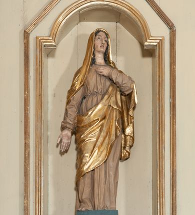 Zdjęcie nr 15: Ołtarz architektoniczny, jednokondygnacyjny, trójosiowy, ze zwieńczeniem. Ołtarz prostopadłościenny, z drewnianą mensą. Antepedium w formie haftowanej tkaniny zdobionej po środku motywem zwieńczonego krzyżem monogramu maryjnego z koroną, ujętego dwunastoma gwiazdami, natomiast w dolnych narożach gałązkami z kwiatami białych lilii.  Tabernakulum z tronem wystawienia w formie jednokondygnacyjnej, trójosiowej bryły architektonicznej, osadzonej na wysokim cokole. Tabernakulum wbudowane w osi środkowej cokołu, drzwiczki prostokątne, zdobione płaskorzeźbionym motywem kielicha eucharystycznego. Flankowane po skrajnych bokach parą pilastrów osie boczne całej struktury są wklęsłe i cofnięte względem osi środkowej ujętej parą kolumienek. Podpory wspierają wyłamujące się ponad nimi belkowanie z gzymsem po środku w formie łuku koszowego. Część środkowa w formie niszy zamkniętej motywem konchy. W zwieńczeniu figurka baranka paschalnego ujętego promienistą glorią, ukazanego na owalnej podstawie osadzonej na 4 wolutowych kabłąkach. Po bokach para figur adorujących aniołów. Cokół zdobiony motywami uskrzydlonych główek anielskich, rocaille’i oraz gałązek z kiściami winnej latorośli. Pola osi bocznych wypełnia para podwieszeń w formie gałązek różanych, partie fryzu pojedyncze rozetki. Struktura tabernakulum i tronu wystawienia jest złocona i srebrzona.   Retabulum osadzone na zdwojonym, zryzalitowanym uskokowo po bokach cokole. Część środkowa ujęta dwoma parami kolumn – wewnętrzne ustawione na tle pary pilastrów, zewnętrzne na tle pary filarów. Podpory dźwigają wyłamujące się ponad nimi belkowanie przerwane w partii architrawu i fryzu. Zwieńczenie ujęte parą spływów wolutowych, zamknięte parą stykających się wolut. Po bokach przerwany, wolutowy przyczółek. W dolnych polach osi bocznych retabulum para półkoliście zamkniętych bramek, ujętych prostą arkadą. Pola osi oddzielone parami stykających się wolut zwieńczonych konchą. W górnej strefie para płytkich niszy zamkniętych półkolistymi łukami nadwieszonymi i dodatkowo ujęta prostym, trójkątnie zamkniętym obramieniem. Osie zwieńczone trzema fragmentami gzymsu oddzielonymi parami wklęsłości.  Pola cokołu osi środkowej retabulum wypełnione ornamentyką roślinną. Część środkowa ujęta parą ażurowych, ornamentalnych  uszaków. Po bokach pola para zwisów w postaci gałązek różanych, w górnych narożach para rozetek. Pola fryzu zdobione rozetami. Pole zwieńczenia w górnej partii dekorowane parą ulistnionych gałązek, po bokach parą kampanuli, dołem w narożach para dużych rozet, pośrodku wić roślinna. Na spływach wolutowych para płomienistych wazonów, a na przyczółku para figur aniołów. Nadłucza bramek osi bocznych zdobione ornamentem roślinnym, nisze ujęte parą stylizowanych kandelabrów, wyżej para rombów z rozetami, pośrodku gwiazda. Na gzymsach wieńczących płomieniste wazony. W polu środkowym obraz w kształcie stojącego prostokąta zamkniętego odcinkowym łukiem nadwieszonym z uskokami, z wizerunkiem Madonny z Dzieciątkiem. W zwieńczeniu obraz w kształcie stojącego prostokąta zamkniętego odcinkowym łukiem nadwieszonym z uskokami, z wizerunkiem Chrystusa Króla. Nisze osi bocznych wypełniają figury, po prawej stronie św. Barbary, po lewej nieznanej świętej. Struktura w kolorze beżowym, detal architektoniczny i ornamentyka złocone.     