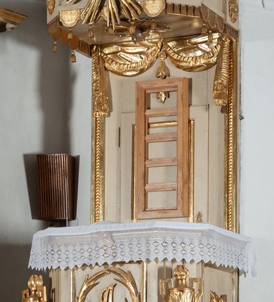 Zdjęcie nr 2: Ambona przyścienna, wisząca, z wejściem od strony zaplecka. Kosz jest na planie czworoboku o parze ściętych naroży artykułowanych samodzielnymi i wysuniętymi do przodu oraz wspartymi na wolutach cokolikami z ustawionymi na nich płonącymi wazonami. Pola ścianek kosza wypełniają płyciny w formie stojących prostokątów, boczne zdobi motyw rozety, środkową monogram S(?) ujęty parą związanych gałązek laurowych. Dół kosza obiega fryz. Podwieszenie kosza, o wklęsłych powierzchniach, zakończone jest motywem złoconej szyszki. Parapet ambony jest w formie gzymsu wieńczącego. W polu zaplecka ujętym motywem podwieszonej kotary z kampanullą pośrodku znajdują się prowadzące na ambonę drzwi. Baldachim jest na rzucie czworoboku o parze ściętych naroży ujętych niewielkimi pilastrami zdobionymi rozetami. Dolną krawędź obiega motyw lambrekinu naprzemiennie z motywem guttów. Czoło baldachimu zdobi promienista gloria z trójkątem z okiem Opatrzności Bożej, natomiast podniebie motyw gołębicy Ducha Świętego na tle promienistej glorii. Zwieńczenie baldachimu ma formę kolumny o ściętym, kanelurowanym trzonie z osadzonym na nim płonącym wazonem. Struktura ambony utrzymana jest w kolorze beżu, detal architektoniczny i ornamentyka są złocone i srebrzone.  