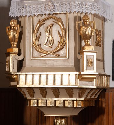 Zdjęcie nr 4: Ambona przyścienna, wisząca, z wejściem od strony zaplecka. Kosz jest na planie czworoboku o parze ściętych naroży artykułowanych samodzielnymi i wysuniętymi do przodu oraz wspartymi na wolutach cokolikami z ustawionymi na nich płonącymi wazonami. Pola ścianek kosza wypełniają płyciny w formie stojących prostokątów, boczne zdobi motyw rozety, środkową monogram S(?) ujęty parą związanych gałązek laurowych. Dół kosza obiega fryz. Podwieszenie kosza, o wklęsłych powierzchniach, zakończone jest motywem złoconej szyszki. Parapet ambony jest w formie gzymsu wieńczącego. W polu zaplecka ujętym motywem podwieszonej kotary z kampanullą pośrodku znajdują się prowadzące na ambonę drzwi. Baldachim jest na rzucie czworoboku o parze ściętych naroży ujętych niewielkimi pilastrami zdobionymi rozetami. Dolną krawędź obiega motyw lambrekinu naprzemiennie z motywem guttów. Czoło baldachimu zdobi promienista gloria z trójkątem z okiem Opatrzności Bożej, natomiast podniebie motyw gołębicy Ducha Świętego na tle promienistej glorii. Zwieńczenie baldachimu ma formę kolumny o ściętym, kanelurowanym trzonie z osadzonym na nim płonącym wazonem. Struktura ambony utrzymana jest w kolorze beżu, detal architektoniczny i ornamentyka są złocone i srebrzone.  