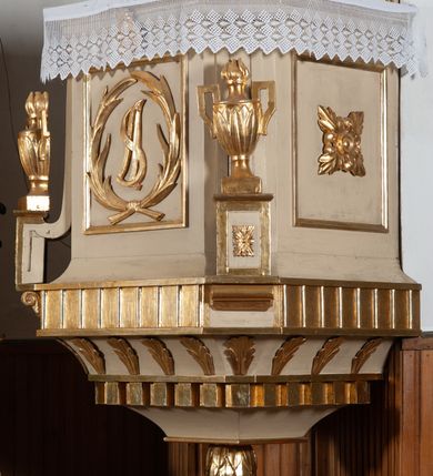 Zdjęcie nr 3: Ambona przyścienna, wisząca, z wejściem od strony zaplecka. Kosz jest na planie czworoboku o parze ściętych naroży artykułowanych samodzielnymi i wysuniętymi do przodu oraz wspartymi na wolutach cokolikami z ustawionymi na nich płonącymi wazonami. Pola ścianek kosza wypełniają płyciny w formie stojących prostokątów, boczne zdobi motyw rozety, środkową monogram S(?) ujęty parą związanych gałązek laurowych. Dół kosza obiega fryz. Podwieszenie kosza, o wklęsłych powierzchniach, zakończone jest motywem złoconej szyszki. Parapet ambony jest w formie gzymsu wieńczącego. W polu zaplecka ujętym motywem podwieszonej kotary z kampanullą pośrodku znajdują się prowadzące na ambonę drzwi. Baldachim jest na rzucie czworoboku o parze ściętych naroży ujętych niewielkimi pilastrami zdobionymi rozetami. Dolną krawędź obiega motyw lambrekinu naprzemiennie z motywem guttów. Czoło baldachimu zdobi promienista gloria z trójkątem z okiem Opatrzności Bożej, natomiast podniebie motyw gołębicy Ducha Świętego na tle promienistej glorii. Zwieńczenie baldachimu ma formę kolumny o ściętym, kanelurowanym trzonie z osadzonym na nim płonącym wazonem. Struktura ambony utrzymana jest w kolorze beżu, detal architektoniczny i ornamentyka są złocone i srebrzone.  