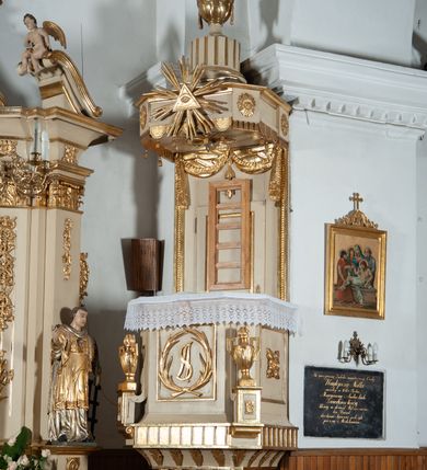 Zdjęcie nr 1: Ambona przyścienna, wisząca, z wejściem od strony zaplecka. Kosz jest na planie czworoboku o parze ściętych naroży artykułowanych samodzielnymi i wysuniętymi do przodu oraz wspartymi na wolutach cokolikami z ustawionymi na nich płonącymi wazonami. Pola ścianek kosza wypełniają płyciny w formie stojących prostokątów, boczne zdobi motyw rozety, środkową monogram S(?) ujęty parą związanych gałązek laurowych. Dół kosza obiega fryz. Podwieszenie kosza, o wklęsłych powierzchniach, zakończone jest motywem złoconej szyszki. Parapet ambony jest w formie gzymsu wieńczącego. W polu zaplecka ujętym motywem podwieszonej kotary z kampanullą pośrodku znajdują się prowadzące na ambonę drzwi. Baldachim jest na rzucie czworoboku o parze ściętych naroży ujętych niewielkimi pilastrami zdobionymi rozetami. Dolną krawędź obiega motyw lambrekinu naprzemiennie z motywem guttów. Czoło baldachimu zdobi promienista gloria z trójkątem z okiem Opatrzności Bożej, natomiast podniebie motyw gołębicy Ducha Świętego na tle promienistej glorii. Zwieńczenie baldachimu ma formę kolumny o ściętym, kanelurowanym trzonie z osadzonym na nim płonącym wazonem. Struktura ambony utrzymana jest w kolorze beżu, detal architektoniczny i ornamentyka są złocone i srebrzone.  