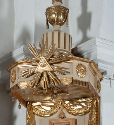 Zdjęcie nr 5: Ambona przyścienna, wisząca, z wejściem od strony zaplecka. Kosz jest na planie czworoboku o parze ściętych naroży artykułowanych samodzielnymi i wysuniętymi do przodu oraz wspartymi na wolutach cokolikami z ustawionymi na nich płonącymi wazonami. Pola ścianek kosza wypełniają płyciny w formie stojących prostokątów, boczne zdobi motyw rozety, środkową monogram S(?) ujęty parą związanych gałązek laurowych. Dół kosza obiega fryz. Podwieszenie kosza, o wklęsłych powierzchniach, zakończone jest motywem złoconej szyszki. Parapet ambony jest w formie gzymsu wieńczącego. W polu zaplecka ujętym motywem podwieszonej kotary z kampanullą pośrodku znajdują się prowadzące na ambonę drzwi. Baldachim jest na rzucie czworoboku o parze ściętych naroży ujętych niewielkimi pilastrami zdobionymi rozetami. Dolną krawędź obiega motyw lambrekinu naprzemiennie z motywem guttów. Czoło baldachimu zdobi promienista gloria z trójkątem z okiem Opatrzności Bożej, natomiast podniebie motyw gołębicy Ducha Świętego na tle promienistej glorii. Zwieńczenie baldachimu ma formę kolumny o ściętym, kanelurowanym trzonie z osadzonym na nim płonącym wazonem. Struktura ambony utrzymana jest w kolorze beżu, detal architektoniczny i ornamentyka są złocone i srebrzone.  