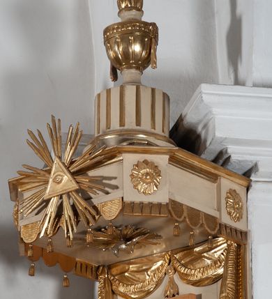 Zdjęcie nr 6: Ambona przyścienna, wisząca, z wejściem od strony zaplecka. Kosz jest na planie czworoboku o parze ściętych naroży artykułowanych samodzielnymi i wysuniętymi do przodu oraz wspartymi na wolutach cokolikami z ustawionymi na nich płonącymi wazonami. Pola ścianek kosza wypełniają płyciny w formie stojących prostokątów, boczne zdobi motyw rozety, środkową monogram S(?) ujęty parą związanych gałązek laurowych. Dół kosza obiega fryz. Podwieszenie kosza, o wklęsłych powierzchniach, zakończone jest motywem złoconej szyszki. Parapet ambony jest w formie gzymsu wieńczącego. W polu zaplecka ujętym motywem podwieszonej kotary z kampanullą pośrodku znajdują się prowadzące na ambonę drzwi. Baldachim jest na rzucie czworoboku o parze ściętych naroży ujętych niewielkimi pilastrami zdobionymi rozetami. Dolną krawędź obiega motyw lambrekinu naprzemiennie z motywem guttów. Czoło baldachimu zdobi promienista gloria z trójkątem z okiem Opatrzności Bożej, natomiast podniebie motyw gołębicy Ducha Świętego na tle promienistej glorii. Zwieńczenie baldachimu ma formę kolumny o ściętym, kanelurowanym trzonie z osadzonym na nim płonącym wazonem. Struktura ambony utrzymana jest w kolorze beżu, detal architektoniczny i ornamentyka są złocone i srebrzone.  