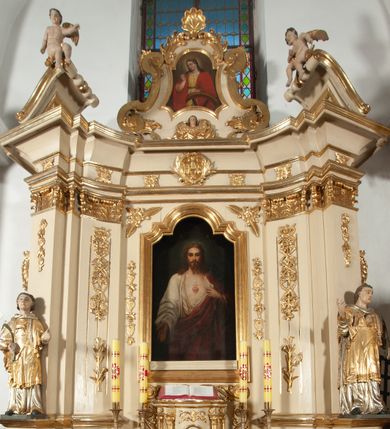 Zdjęcie nr 23: Ołtarz architektoniczny, jednokondygnacyjny, jednoosiowy ze zwieńczeniem. Ołtarz prostopadłościenny, z drewnianą mensą, z antepedium ujętym po bokach parą konsoli w formie wolut. Pole antepedium w postaci prostokątnej płyciny z płaskorzeźbionym motywem gorejącego serca pośrodku, ujętego promienistą glorią. Na mensie tabernakulum w formie prostopadłościennej szafki o wypukłej, przedniej  ściance ujętej dwoma parami osadzonych na niewielkich cokołach kolumienek wspierających niepełne, wyłamujące się ponad nimi belkowanie. Drzwiczki w kształcie stojącego prostokąta zamkniętego półkolistym łukiem nadwieszonym, zdobione motywem płaskorzeźbionego kielicha eucharystycznego. Struktura zdobiona motywami rozet i wici roślinnej. Retabulum osadzone na niewysokim, zryzalitowanym po bokach cokole. Część środkową nastawy ujmuje para ustawionych skośnie  filarów wspierających wyłamujące się ponad nimi belkowanie. Zamknięte półkoliście zwieńczenie ujmuje para spływów wolutowych oraz flankuje przerwany, wolutowy przyczółek. Pola wolutowych konsoli zdobi para liści akantu, cokół retabulum para stylizowanych słoneczników i S-kształtnych wici. Zewnętrzne pola trzonów filarów zdobią w górnych partiach ukwiecone gałązki, wewnętrzne motyw podwieszeń skomponowanych z zamkniętej korony, krzyżujących się rogów obfitości i krzyża z kotwicą, oraz kokard, u dołu para gałązek. Boki pola środkowego z motywem wici winnego grona, u góry w narożach para ulistnionych rozet. Fryz zdobiony rozetami, pośrodku kartusz w monogramem imienia Jezus ujętym parą gałązek laurowych. Retabulum flankowane parą ażurowych uszaków rocaille’owych. W dolnej partii zwieńczenia uskrzydlona główka anielska, szczyt w formie konchy ujętej po bokach parą rocaille’i. Na przerwanym przyczółku para figur aniołków. Pole części środkowej w kształcie stojącego prostokąta zamkniętego półkolistym łukiem nadwieszonym z uskokami, ujęte profilowaną, złoconą ramą, w środku wizerunek Najświętszego Serca Jezusa. Po bokach, na tle filarów para figur, po lewej św. Szczepan, po prawej św. Wawrzyniec. W zwieńczeniu obraz w kształcie trapezu o wklęsłych ramionach, podstawie i dolnych narożach, zamknięty półkolistym łukiem nadwieszonym, ujęty profilowaną, złoconą ramą, z wizerunkiem św. Katarzyny Aleksandryjskiej. 
Struktura w kolorze kremowym, detal architektoniczny i ornamentyka złocone.    
