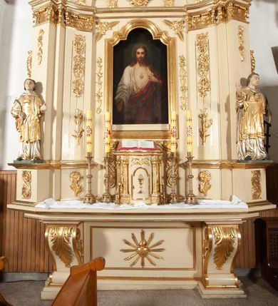 Zdjęcie nr 13: Ołtarz architektoniczny, jednokondygnacyjny, jednoosiowy ze zwieńczeniem. Ołtarz prostopadłościenny, z drewnianą mensą, z antepedium ujętym po bokach parą konsoli w formie wolut. Pole antepedium w postaci prostokątnej płyciny z płaskorzeźbionym motywem gorejącego serca pośrodku, ujętego promienistą glorią. Na mensie tabernakulum w formie prostopadłościennej szafki o wypukłej, przedniej  ściance ujętej dwoma parami osadzonych na niewielkich cokołach kolumienek wspierających niepełne, wyłamujące się ponad nimi belkowanie. Drzwiczki w kształcie stojącego prostokąta zamkniętego półkolistym łukiem nadwieszonym, zdobione motywem płaskorzeźbionego kielicha eucharystycznego. Struktura zdobiona motywami rozet i wici roślinnej. Retabulum osadzone na niewysokim, zryzalitowanym po bokach cokole. Część środkową nastawy ujmuje para ustawionych skośnie  filarów wspierających wyłamujące się ponad nimi belkowanie. Zamknięte półkoliście zwieńczenie ujmuje para spływów wolutowych oraz flankuje przerwany, wolutowy przyczółek. Pola wolutowych konsoli zdobi para liści akantu, cokół retabulum para stylizowanych słoneczników i S-kształtnych wici. Zewnętrzne pola trzonów filarów zdobią w górnych partiach ukwiecone gałązki, wewnętrzne motyw podwieszeń skomponowanych z zamkniętej korony, krzyżujących się rogów obfitości i krzyża z kotwicą, oraz kokard, u dołu para gałązek. Boki pola środkowego z motywem wici winnego grona, u góry w narożach para ulistnionych rozet. Fryz zdobiony rozetami, pośrodku kartusz w monogramem imienia Jezus ujętym parą gałązek laurowych. Retabulum flankowane parą ażurowych uszaków rocaille’owych. W dolnej partii zwieńczenia uskrzydlona główka anielska, szczyt w formie konchy ujętej po bokach parą rocaille’i. Na przerwanym przyczółku para figur aniołków. Pole części środkowej w kształcie stojącego prostokąta zamkniętego półkolistym łukiem nadwieszonym z uskokami, ujęte profilowaną, złoconą ramą, w środku wizerunek Najświętszego Serca Jezusa. Po bokach, na tle filarów para figur, po lewej św. Szczepan, po prawej św. Wawrzyniec. W zwieńczeniu obraz w kształcie trapezu o wklęsłych ramionach, podstawie i dolnych narożach, zamknięty półkolistym łukiem nadwieszonym, ujęty profilowaną, złoconą ramą, z wizerunkiem św. Katarzyny Aleksandryjskiej. 
Struktura w kolorze kremowym, detal architektoniczny i ornamentyka złocone.    
