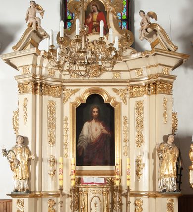 Zdjęcie nr 2: Ołtarz architektoniczny, jednokondygnacyjny, jednoosiowy ze zwieńczeniem. Ołtarz prostopadłościenny, z drewnianą mensą, z antepedium ujętym po bokach parą konsoli w formie wolut. Pole antepedium w postaci prostokątnej płyciny z płaskorzeźbionym motywem gorejącego serca pośrodku, ujętego promienistą glorią. Na mensie tabernakulum w formie prostopadłościennej szafki o wypukłej, przedniej  ściance ujętej dwoma parami osadzonych na niewielkich cokołach kolumienek wspierających niepełne, wyłamujące się ponad nimi belkowanie. Drzwiczki w kształcie stojącego prostokąta zamkniętego półkolistym łukiem nadwieszonym, zdobione motywem płaskorzeźbionego kielicha eucharystycznego. Struktura zdobiona motywami rozet i wici roślinnej. Retabulum osadzone na niewysokim, zryzalitowanym po bokach cokole. Część środkową nastawy ujmuje para ustawionych skośnie  filarów wspierających wyłamujące się ponad nimi belkowanie. Zamknięte półkoliście zwieńczenie ujmuje para spływów wolutowych oraz flankuje przerwany, wolutowy przyczółek. Pola wolutowych konsoli zdobi para liści akantu, cokół retabulum para stylizowanych słoneczników i S-kształtnych wici. Zewnętrzne pola trzonów filarów zdobią w górnych partiach ukwiecone gałązki, wewnętrzne motyw podwieszeń skomponowanych z zamkniętej korony, krzyżujących się rogów obfitości i krzyża z kotwicą, oraz kokard, u dołu para gałązek. Boki pola środkowego z motywem wici winnego grona, u góry w narożach para ulistnionych rozet. Fryz zdobiony rozetami, pośrodku kartusz w monogramem imienia Jezus ujętym parą gałązek laurowych. Retabulum flankowane parą ażurowych uszaków rocaille’owych. W dolnej partii zwieńczenia uskrzydlona główka anielska, szczyt w formie konchy ujętej po bokach parą rocaille’i. Na przerwanym przyczółku para figur aniołków. Pole części środkowej w kształcie stojącego prostokąta zamkniętego półkolistym łukiem nadwieszonym z uskokami, ujęte profilowaną, złoconą ramą, w środku wizerunek Najświętszego Serca Jezusa. Po bokach, na tle filarów para figur, po lewej św. Szczepan, po prawej św. Wawrzyniec. W zwieńczeniu obraz w kształcie trapezu o wklęsłych ramionach, podstawie i dolnych narożach, zamknięty półkolistym łukiem nadwieszonym, ujęty profilowaną, złoconą ramą, z wizerunkiem św. Katarzyny Aleksandryjskiej. 
Struktura w kolorze kremowym, detal architektoniczny i ornamentyka złocone.    
