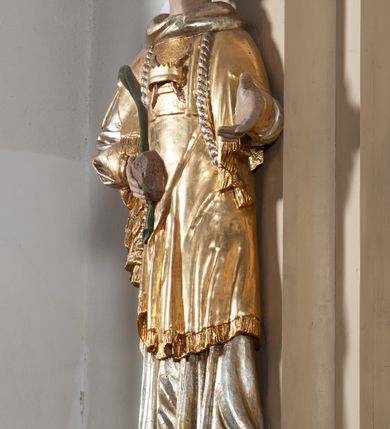 Zdjęcie nr 3: Całopostaciowa, pełnoplastyczna figura św. Szczepana stojącego w lekkim kontrapoście na niewysokiej podstawie, zwróconego w trzech czwartych w lewo. Podniesioną głowę charakteryzuje owalna twarz o młodzieńczych  rysach z niskim czołem, wysoko uniesionymi brwiami, dużymi oczami długim nosem i kształtnymi ustami w lekkim uśmiechu. Włosy krótkie, sięgające szyi, z krótką grzywką, odsłaniające uszy. Święty ubrany w srebrzoną, silnie fałdowaną albę obszytą ząbkowaną lamówką oraz złoconą, obszytą frędzlą dalmatykę z motywem korony z parą gałązek na piersiach oraz parą grubych sznurów zakończonych chwostami. Na nogach czarne buty. Lewa, zgięta w łokciu ręka wyciągnięta przed siebie z dłonią skierowaną w dół, prawą podtrzymuje kamień i gałązkę palmową.       