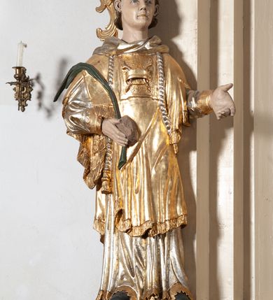 Zdjęcie nr 4: Całopostaciowa, pełnoplastyczna figura św. Szczepana stojącego w lekkim kontrapoście na niewysokiej podstawie, zwróconego w trzech czwartych w lewo. Podniesioną głowę charakteryzuje owalna twarz o młodzieńczych  rysach z niskim czołem, wysoko uniesionymi brwiami, dużymi oczami długim nosem i kształtnymi ustami w lekkim uśmiechu. Włosy krótkie, sięgające szyi, z krótką grzywką, odsłaniające uszy. Święty ubrany w srebrzoną, silnie fałdowaną albę obszytą ząbkowaną lamówką oraz złoconą, obszytą frędzlą dalmatykę z motywem korony z parą gałązek na piersiach oraz parą grubych sznurów zakończonych chwostami. Na nogach czarne buty. Lewa, zgięta w łokciu ręka wyciągnięta przed siebie z dłonią skierowaną w dół, prawą podtrzymuje kamień i gałązkę palmową.       