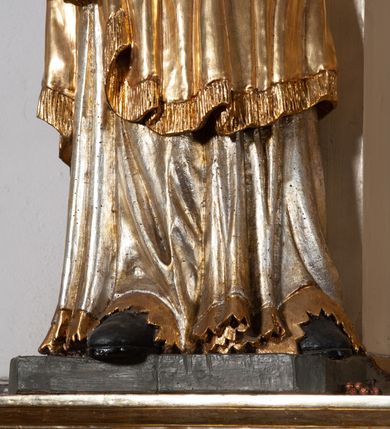 Zdjęcie nr 7: Całopostaciowa, pełnoplastyczna figura św. Szczepana stojącego w lekkim kontrapoście na niewysokiej podstawie, zwróconego w trzech czwartych w lewo. Podniesioną głowę charakteryzuje owalna twarz o młodzieńczych  rysach z niskim czołem, wysoko uniesionymi brwiami, dużymi oczami długim nosem i kształtnymi ustami w lekkim uśmiechu. Włosy krótkie, sięgające szyi, z krótką grzywką, odsłaniające uszy. Święty ubrany w srebrzoną, silnie fałdowaną albę obszytą ząbkowaną lamówką oraz złoconą, obszytą frędzlą dalmatykę z motywem korony z parą gałązek na piersiach oraz parą grubych sznurów zakończonych chwostami. Na nogach czarne buty. Lewa, zgięta w łokciu ręka wyciągnięta przed siebie z dłonią skierowaną w dół, prawą podtrzymuje kamień i gałązkę palmową.       