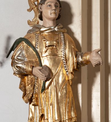 Zdjęcie nr 2: Całopostaciowa, pełnoplastyczna figura św. Szczepana stojącego w lekkim kontrapoście na niewysokiej podstawie, zwróconego w trzech czwartych w lewo. Podniesioną głowę charakteryzuje owalna twarz o młodzieńczych  rysach z niskim czołem, wysoko uniesionymi brwiami, dużymi oczami długim nosem i kształtnymi ustami w lekkim uśmiechu. Włosy krótkie, sięgające szyi, z krótką grzywką, odsłaniające uszy. Święty ubrany w srebrzoną, silnie fałdowaną albę obszytą ząbkowaną lamówką oraz złoconą, obszytą frędzlą dalmatykę z motywem korony z parą gałązek na piersiach oraz parą grubych sznurów zakończonych chwostami. Na nogach czarne buty. Lewa, zgięta w łokciu ręka wyciągnięta przed siebie z dłonią skierowaną w dół, prawą podtrzymuje kamień i gałązkę palmową.       