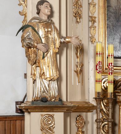 Zdjęcie nr 1: Całopostaciowa, pełnoplastyczna figura św. Szczepana stojącego w lekkim kontrapoście na niewysokiej podstawie, zwróconego w trzech czwartych w lewo. Podniesioną głowę charakteryzuje owalna twarz o młodzieńczych  rysach z niskim czołem, wysoko uniesionymi brwiami, dużymi oczami długim nosem i kształtnymi ustami w lekkim uśmiechu. Włosy krótkie, sięgające szyi, z krótką grzywką, odsłaniające uszy. Święty ubrany w srebrzoną, silnie fałdowaną albę obszytą ząbkowaną lamówką oraz złoconą, obszytą frędzlą dalmatykę z motywem korony z parą gałązek na piersiach oraz parą grubych sznurów zakończonych chwostami. Na nogach czarne buty. Lewa, zgięta w łokciu ręka wyciągnięta przed siebie z dłonią skierowaną w dół, prawą podtrzymuje kamień i gałązkę palmową.       