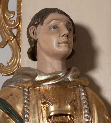 Zdjęcie nr 6: Całopostaciowa, pełnoplastyczna figura św. Szczepana stojącego w lekkim kontrapoście na niewysokiej podstawie, zwróconego w trzech czwartych w lewo. Podniesioną głowę charakteryzuje owalna twarz o młodzieńczych  rysach z niskim czołem, wysoko uniesionymi brwiami, dużymi oczami długim nosem i kształtnymi ustami w lekkim uśmiechu. Włosy krótkie, sięgające szyi, z krótką grzywką, odsłaniające uszy. Święty ubrany w srebrzoną, silnie fałdowaną albę obszytą ząbkowaną lamówką oraz złoconą, obszytą frędzlą dalmatykę z motywem korony z parą gałązek na piersiach oraz parą grubych sznurów zakończonych chwostami. Na nogach czarne buty. Lewa, zgięta w łokciu ręka wyciągnięta przed siebie z dłonią skierowaną w dół, prawą podtrzymuje kamień i gałązkę palmową.       