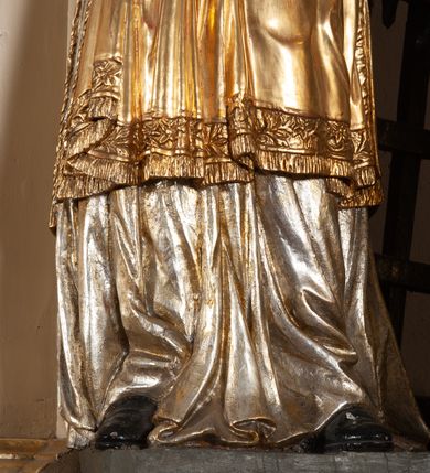 Zdjęcie nr 7: Całopostaciowa, pełnoplastyczna figura św. Wawrzyńca stojącego w kontrapoście na niewysokiej podstawie, zwróconego na wprost. Skierowaną nieznacznie w lewo głowę charakteryzuje owalna twarz o młodzieńczych  rysach z niskim czołem, wysoko uniesionymi brwiami, dużymi oczami, długim nosem i wąskimi ustami ujętymi bruzdami. Włosy krótkie, zaczesane do tyłu, odsłaniające uszy. Święty ubrany jest w srebrzoną, silnie fałdowaną albę i złoconą, obszytą glonem i frędzlą dalmatykę z motywem uskrzydlonej główki anielskiej na piersiach. Na nogach ma czarne buty. Prawa, zgięta w łokciu ręka uniesiona do góry z wyprostowanym palcem wskazującym, lewa opuszczona w dół, z gałązką palmową w dłoni.      

