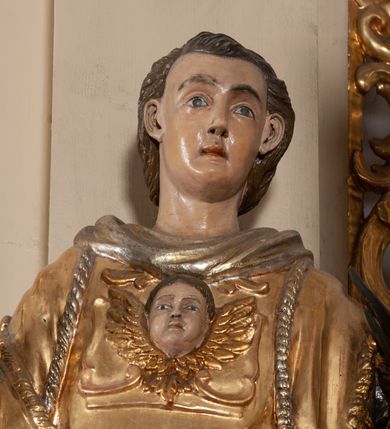 Zdjęcie nr 3: Całopostaciowa, pełnoplastyczna figura św. Wawrzyńca stojącego w kontrapoście na niewysokiej podstawie, zwróconego na wprost. Skierowaną nieznacznie w lewo głowę charakteryzuje owalna twarz o młodzieńczych  rysach z niskim czołem, wysoko uniesionymi brwiami, dużymi oczami, długim nosem i wąskimi ustami ujętymi bruzdami. Włosy krótkie, zaczesane do tyłu, odsłaniające uszy. Święty ubrany jest w srebrzoną, silnie fałdowaną albę i złoconą, obszytą glonem i frędzlą dalmatykę z motywem uskrzydlonej główki anielskiej na piersiach. Na nogach ma czarne buty. Prawa, zgięta w łokciu ręka uniesiona do góry z wyprostowanym palcem wskazującym, lewa opuszczona w dół, z gałązką palmową w dłoni.      

