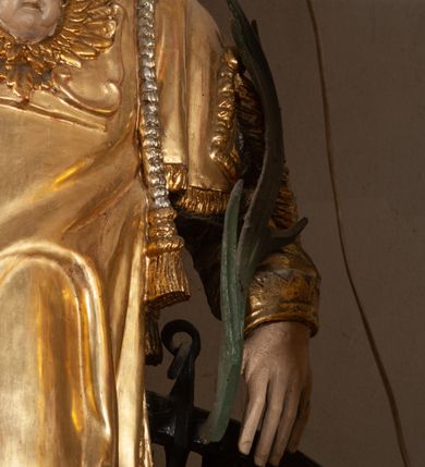 Zdjęcie nr 5: Całopostaciowa, pełnoplastyczna figura św. Wawrzyńca stojącego w kontrapoście na niewysokiej podstawie, zwróconego na wprost. Skierowaną nieznacznie w lewo głowę charakteryzuje owalna twarz o młodzieńczych  rysach z niskim czołem, wysoko uniesionymi brwiami, dużymi oczami, długim nosem i wąskimi ustami ujętymi bruzdami. Włosy krótkie, zaczesane do tyłu, odsłaniające uszy. Święty ubrany jest w srebrzoną, silnie fałdowaną albę i złoconą, obszytą glonem i frędzlą dalmatykę z motywem uskrzydlonej główki anielskiej na piersiach. Na nogach ma czarne buty. Prawa, zgięta w łokciu ręka uniesiona do góry z wyprostowanym palcem wskazującym, lewa opuszczona w dół, z gałązką palmową w dłoni.      

