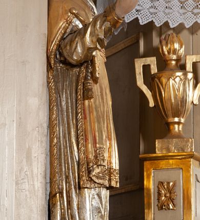 Zdjęcie nr 4: Całopostaciowa, pełnoplastyczna figura św. Wawrzyńca stojącego w kontrapoście na niewysokiej podstawie, zwróconego na wprost. Skierowaną nieznacznie w lewo głowę charakteryzuje owalna twarz o młodzieńczych  rysach z niskim czołem, wysoko uniesionymi brwiami, dużymi oczami, długim nosem i wąskimi ustami ujętymi bruzdami. Włosy krótkie, zaczesane do tyłu, odsłaniające uszy. Święty ubrany jest w srebrzoną, silnie fałdowaną albę i złoconą, obszytą glonem i frędzlą dalmatykę z motywem uskrzydlonej główki anielskiej na piersiach. Na nogach ma czarne buty. Prawa, zgięta w łokciu ręka uniesiona do góry z wyprostowanym palcem wskazującym, lewa opuszczona w dół, z gałązką palmową w dłoni.      

