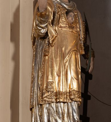 Zdjęcie nr 2: Całopostaciowa, pełnoplastyczna figura św. Wawrzyńca stojącego w kontrapoście na niewysokiej podstawie, zwróconego na wprost. Skierowaną nieznacznie w lewo głowę charakteryzuje owalna twarz o młodzieńczych  rysach z niskim czołem, wysoko uniesionymi brwiami, dużymi oczami, długim nosem i wąskimi ustami ujętymi bruzdami. Włosy krótkie, zaczesane do tyłu, odsłaniające uszy. Święty ubrany jest w srebrzoną, silnie fałdowaną albę i złoconą, obszytą glonem i frędzlą dalmatykę z motywem uskrzydlonej główki anielskiej na piersiach. Na nogach ma czarne buty. Prawa, zgięta w łokciu ręka uniesiona do góry z wyprostowanym palcem wskazującym, lewa opuszczona w dół, z gałązką palmową w dłoni.      

