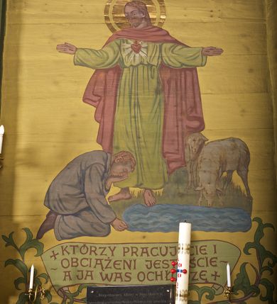 Zdjęcie nr 2: Ściany kościoła dekorują przedstawienia świętych: św. Stanisław w stroju pontyfikalnym z pastorałem, wskrzeszający Piotrowina. Poniżej na banderoli napis „BŁOGOSŁAWIENI KTÓRZY ŁAKNĄ / I PRAGNĄ SPRAWIEDLIWOŚCI”; św. Jadwiga w koronie i płaszczu podszytym gronostajem, trzymająca skrzyneczkę z klejnotami oraz krzyż z herbami Polski i Litwy. Na banderoli napis „BŁOGOSŁAWIENI / POKÓJ CZYNIĄCY”; św. Jan Kanty błogosławiący dziewczynę z dzbanem podpisany „BŁOGOSŁAWIENI / MIŁOSIERNI”; św. w habicie, z krzyżem w ręce i w asyście dwóch aniołów - jednego z palmą męczeństwa, drugiego z płonącą pochodnią, pod nimi napis „BŁOGOSŁAWIENI KTÓRZY / CIERPIĄ PRZEŚLADOWANIA DLA SPRAWIEDLIWOŚCI”; Chrystus z sercem gorejącym. Po jego prawej stronie klęczący mężczyzna, po lewej dwie owieczki nad strumieniem. Scena podpisana: „KTÓRZY PRACUJECIE I / OBCIĄŻENI JESTEŚCIE / A JA WAS OCHŁODZĘ”; archanioł Gabriel z lilią powyżej napisu  „ZDROWAŚ MARIO”; archanioł Michał z trybularzem, ponad napisem „KTÓŻ JAK BÓG!”; św. Franciszek na tle przyrody. Jest ubrany w habit franciszkański, na głowie ma kaptur. Przy prawej nodze świętego wilk, w jeziorze ryby, na niebie ptaki. Na banderoli napis „BŁOGOSŁAWIENI UBODZY DUCHEM / ALBOWIEM TAKOWYCH JEST / KRÓLESTWO NIEBIESKIE”; św. Izydor Oracz ukazany w towarzystwie anioła orającego na polu na tle dzwonnicy. Poniżej napis „BŁOGOSŁAWIENI CISI / ALBOWIEM ONI / POSIĄDĄ ZIEMIĘ”. Poniżej postaci świętych fryz z przedstawieniem 14 stacji drogi krzyżowej na błękitnym tle. Na stropie nawy i prezbiterium, na błękitnym tle, wśród kwiatowego ornamentu na środku ukazany Chrystus Ukrzyżowany ponad kielichem, gołębica Ducha Świętego i koronowana Maria. Po ich obu stronach stoją święci mężowie i niewiasty z atrybutami. W nawie w kolejności do środka: św. Wojciech (?), św. Jacek z figurą Matki Boskiej i monstrancją, klęczący św. Kazimierz w stroju książęcym z lilią, św. Agnieszka z barankiem i św. Karolina Kózkówna z różańcem, klęcząca św. Rozalia z różami, św. Klara z cyborium, św. Weronika z chustą z wizerunkiem Chrystusa, klęcząca św. Cecylia z lirą, św. Józef z lilią, św. Paweł z mieczem, klęczący św. Jan Chrzciciel z krzyżem. W prezbiterium przedstawiono 12 apostołów, niektórzy z atrybutmi, które ich jednoznacznie identyfilują, np. św. Andrzej z krzyżem w kształcie litery X, inni z księgami lub zwojami.  