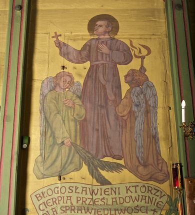 Zdjęcie nr 4: Ściany kościoła dekorują przedstawienia świętych: św. Stanisław w stroju pontyfikalnym z pastorałem, wskrzeszający Piotrowina. Poniżej na banderoli napis „BŁOGOSŁAWIENI KTÓRZY ŁAKNĄ / I PRAGNĄ SPRAWIEDLIWOŚCI”; św. Jadwiga w koronie i płaszczu podszytym gronostajem, trzymająca skrzyneczkę z klejnotami oraz krzyż z herbami Polski i Litwy. Na banderoli napis „BŁOGOSŁAWIENI / POKÓJ CZYNIĄCY”; św. Jan Kanty błogosławiący dziewczynę z dzbanem podpisany „BŁOGOSŁAWIENI / MIŁOSIERNI”; św. w habicie, z krzyżem w ręce i w asyście dwóch aniołów - jednego z palmą męczeństwa, drugiego z płonącą pochodnią, pod nimi napis „BŁOGOSŁAWIENI KTÓRZY / CIERPIĄ PRZEŚLADOWANIA DLA SPRAWIEDLIWOŚCI”; Chrystus z sercem gorejącym. Po jego prawej stronie klęczący mężczyzna, po lewej dwie owieczki nad strumieniem. Scena podpisana: „KTÓRZY PRACUJECIE I / OBCIĄŻENI JESTEŚCIE / A JA WAS OCHŁODZĘ”; archanioł Gabriel z lilią powyżej napisu  „ZDROWAŚ MARIO”; archanioł Michał z trybularzem, ponad napisem „KTÓŻ JAK BÓG!”; św. Franciszek na tle przyrody. Jest ubrany w habit franciszkański, na głowie ma kaptur. Przy prawej nodze świętego wilk, w jeziorze ryby, na niebie ptaki. Na banderoli napis „BŁOGOSŁAWIENI UBODZY DUCHEM / ALBOWIEM TAKOWYCH JEST / KRÓLESTWO NIEBIESKIE”; św. Izydor Oracz ukazany w towarzystwie anioła orającego na polu na tle dzwonnicy. Poniżej napis „BŁOGOSŁAWIENI CISI / ALBOWIEM ONI / POSIĄDĄ ZIEMIĘ”. Poniżej postaci świętych fryz z przedstawieniem 14 stacji drogi krzyżowej na błękitnym tle. Na stropie nawy i prezbiterium, na błękitnym tle, wśród kwiatowego ornamentu na środku ukazany Chrystus Ukrzyżowany ponad kielichem, gołębica Ducha Świętego i koronowana Maria. Po ich obu stronach stoją święci mężowie i niewiasty z atrybutami. W nawie w kolejności do środka: św. Wojciech (?), św. Jacek z figurą Matki Boskiej i monstrancją, klęczący św. Kazimierz w stroju książęcym z lilią, św. Agnieszka z barankiem i św. Karolina Kózkówna z różańcem, klęcząca św. Rozalia z różami, św. Klara z cyborium, św. Weronika z chustą z wizerunkiem Chrystusa, klęcząca św. Cecylia z lirą, św. Józef z lilią, św. Paweł z mieczem, klęczący św. Jan Chrzciciel z krzyżem. W prezbiterium przedstawiono 12 apostołów, niektórzy z atrybutmi, które ich jednoznacznie identyfilują, np. św. Andrzej z krzyżem w kształcie litery X, inni z księgami lub zwojami.  