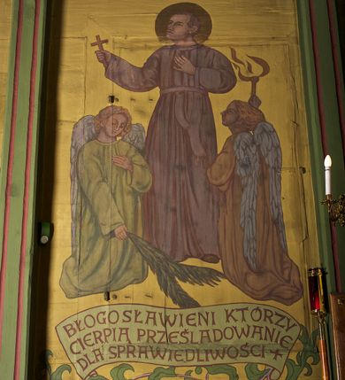 Zdjęcie nr 3: Ściany kościoła dekorują przedstawienia świętych: św. Stanisław w stroju pontyfikalnym z pastorałem, wskrzeszający Piotrowina. Poniżej na banderoli napis „BŁOGOSŁAWIENI KTÓRZY ŁAKNĄ / I PRAGNĄ SPRAWIEDLIWOŚCI”; św. Jadwiga w koronie i płaszczu podszytym gronostajem, trzymająca skrzyneczkę z klejnotami oraz krzyż z herbami Polski i Litwy. Na banderoli napis „BŁOGOSŁAWIENI / POKÓJ CZYNIĄCY”; św. Jan Kanty błogosławiący dziewczynę z dzbanem podpisany „BŁOGOSŁAWIENI / MIŁOSIERNI”; św. w habicie, z krzyżem w ręce i w asyście dwóch aniołów - jednego z palmą męczeństwa, drugiego z płonącą pochodnią, pod nimi napis „BŁOGOSŁAWIENI KTÓRZY / CIERPIĄ PRZEŚLADOWANIA DLA SPRAWIEDLIWOŚCI”; Chrystus z sercem gorejącym. Po jego prawej stronie klęczący mężczyzna, po lewej dwie owieczki nad strumieniem. Scena podpisana: „KTÓRZY PRACUJECIE I / OBCIĄŻENI JESTEŚCIE / A JA WAS OCHŁODZĘ”; archanioł Gabriel z lilią powyżej napisu  „ZDROWAŚ MARIO”; archanioł Michał z trybularzem, ponad napisem „KTÓŻ JAK BÓG!”; św. Franciszek na tle przyrody. Jest ubrany w habit franciszkański, na głowie ma kaptur. Przy prawej nodze świętego wilk, w jeziorze ryby, na niebie ptaki. Na banderoli napis „BŁOGOSŁAWIENI UBODZY DUCHEM / ALBOWIEM TAKOWYCH JEST / KRÓLESTWO NIEBIESKIE”; św. Izydor Oracz ukazany w towarzystwie anioła orającego na polu na tle dzwonnicy. Poniżej napis „BŁOGOSŁAWIENI CISI / ALBOWIEM ONI / POSIĄDĄ ZIEMIĘ”. Poniżej postaci świętych fryz z przedstawieniem 14 stacji drogi krzyżowej na błękitnym tle. Na stropie nawy i prezbiterium, na błękitnym tle, wśród kwiatowego ornamentu na środku ukazany Chrystus Ukrzyżowany ponad kielichem, gołębica Ducha Świętego i koronowana Maria. Po ich obu stronach stoją święci mężowie i niewiasty z atrybutami. W nawie w kolejności do środka: św. Wojciech (?), św. Jacek z figurą Matki Boskiej i monstrancją, klęczący św. Kazimierz w stroju książęcym z lilią, św. Agnieszka z barankiem i św. Karolina Kózkówna z różańcem, klęcząca św. Rozalia z różami, św. Klara z cyborium, św. Weronika z chustą z wizerunkiem Chrystusa, klęcząca św. Cecylia z lirą, św. Józef z lilią, św. Paweł z mieczem, klęczący św. Jan Chrzciciel z krzyżem. W prezbiterium przedstawiono 12 apostołów, niektórzy z atrybutmi, które ich jednoznacznie identyfilują, np. św. Andrzej z krzyżem w kształcie litery X, inni z księgami lub zwojami.  