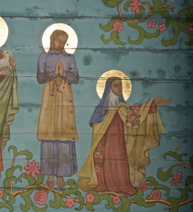 Zdjęcie nr 18: Ściany kościoła dekorują przedstawienia świętych: św. Stanisław w stroju pontyfikalnym z pastorałem, wskrzeszający Piotrowina. Poniżej na banderoli napis „BŁOGOSŁAWIENI KTÓRZY ŁAKNĄ / I PRAGNĄ SPRAWIEDLIWOŚCI”; św. Jadwiga w koronie i płaszczu podszytym gronostajem, trzymająca skrzyneczkę z klejnotami oraz krzyż z herbami Polski i Litwy. Na banderoli napis „BŁOGOSŁAWIENI / POKÓJ CZYNIĄCY”; św. Jan Kanty błogosławiący dziewczynę z dzbanem podpisany „BŁOGOSŁAWIENI / MIŁOSIERNI”; św. w habicie, z krzyżem w ręce i w asyście dwóch aniołów - jednego z palmą męczeństwa, drugiego z płonącą pochodnią, pod nimi napis „BŁOGOSŁAWIENI KTÓRZY / CIERPIĄ PRZEŚLADOWANIA DLA SPRAWIEDLIWOŚCI”; Chrystus z sercem gorejącym. Po jego prawej stronie klęczący mężczyzna, po lewej dwie owieczki nad strumieniem. Scena podpisana: „KTÓRZY PRACUJECIE I / OBCIĄŻENI JESTEŚCIE / A JA WAS OCHŁODZĘ”; archanioł Gabriel z lilią powyżej napisu  „ZDROWAŚ MARIO”; archanioł Michał z trybularzem, ponad napisem „KTÓŻ JAK BÓG!”; św. Franciszek na tle przyrody. Jest ubrany w habit franciszkański, na głowie ma kaptur. Przy prawej nodze świętego wilk, w jeziorze ryby, na niebie ptaki. Na banderoli napis „BŁOGOSŁAWIENI UBODZY DUCHEM / ALBOWIEM TAKOWYCH JEST / KRÓLESTWO NIEBIESKIE”; św. Izydor Oracz ukazany w towarzystwie anioła orającego na polu na tle dzwonnicy. Poniżej napis „BŁOGOSŁAWIENI CISI / ALBOWIEM ONI / POSIĄDĄ ZIEMIĘ”. Poniżej postaci świętych fryz z przedstawieniem 14 stacji drogi krzyżowej na błękitnym tle. Na stropie nawy i prezbiterium, na błękitnym tle, wśród kwiatowego ornamentu na środku ukazany Chrystus Ukrzyżowany ponad kielichem, gołębica Ducha Świętego i koronowana Maria. Po ich obu stronach stoją święci mężowie i niewiasty z atrybutami. W nawie w kolejności do środka: św. Wojciech (?), św. Jacek z figurą Matki Boskiej i monstrancją, klęczący św. Kazimierz w stroju książęcym z lilią, św. Agnieszka z barankiem i św. Karolina Kózkówna z różańcem, klęcząca św. Rozalia z różami, św. Klara z cyborium, św. Weronika z chustą z wizerunkiem Chrystusa, klęcząca św. Cecylia z lirą, św. Józef z lilią, św. Paweł z mieczem, klęczący św. Jan Chrzciciel z krzyżem. W prezbiterium przedstawiono 12 apostołów, niektórzy z atrybutmi, które ich jednoznacznie identyfilują, np. św. Andrzej z krzyżem w kształcie litery X, inni z księgami lub zwojami.  