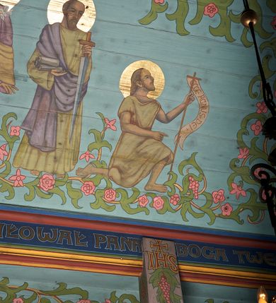Zdjęcie nr 13: Ściany kościoła dekorują przedstawienia świętych: św. Stanisław w stroju pontyfikalnym z pastorałem, wskrzeszający Piotrowina. Poniżej na banderoli napis „BŁOGOSŁAWIENI KTÓRZY ŁAKNĄ / I PRAGNĄ SPRAWIEDLIWOŚCI”; św. Jadwiga w koronie i płaszczu podszytym gronostajem, trzymająca skrzyneczkę z klejnotami oraz krzyż z herbami Polski i Litwy. Na banderoli napis „BŁOGOSŁAWIENI / POKÓJ CZYNIĄCY”; św. Jan Kanty błogosławiący dziewczynę z dzbanem podpisany „BŁOGOSŁAWIENI / MIŁOSIERNI”; św. w habicie, z krzyżem w ręce i w asyście dwóch aniołów - jednego z palmą męczeństwa, drugiego z płonącą pochodnią, pod nimi napis „BŁOGOSŁAWIENI KTÓRZY / CIERPIĄ PRZEŚLADOWANIA DLA SPRAWIEDLIWOŚCI”; Chrystus z sercem gorejącym. Po jego prawej stronie klęczący mężczyzna, po lewej dwie owieczki nad strumieniem. Scena podpisana: „KTÓRZY PRACUJECIE I / OBCIĄŻENI JESTEŚCIE / A JA WAS OCHŁODZĘ”; archanioł Gabriel z lilią powyżej napisu  „ZDROWAŚ MARIO”; archanioł Michał z trybularzem, ponad napisem „KTÓŻ JAK BÓG!”; św. Franciszek na tle przyrody. Jest ubrany w habit franciszkański, na głowie ma kaptur. Przy prawej nodze świętego wilk, w jeziorze ryby, na niebie ptaki. Na banderoli napis „BŁOGOSŁAWIENI UBODZY DUCHEM / ALBOWIEM TAKOWYCH JEST / KRÓLESTWO NIEBIESKIE”; św. Izydor Oracz ukazany w towarzystwie anioła orającego na polu na tle dzwonnicy. Poniżej napis „BŁOGOSŁAWIENI CISI / ALBOWIEM ONI / POSIĄDĄ ZIEMIĘ”. Poniżej postaci świętych fryz z przedstawieniem 14 stacji drogi krzyżowej na błękitnym tle. Na stropie nawy i prezbiterium, na błękitnym tle, wśród kwiatowego ornamentu na środku ukazany Chrystus Ukrzyżowany ponad kielichem, gołębica Ducha Świętego i koronowana Maria. Po ich obu stronach stoją święci mężowie i niewiasty z atrybutami. W nawie w kolejności do środka: św. Wojciech (?), św. Jacek z figurą Matki Boskiej i monstrancją, klęczący św. Kazimierz w stroju książęcym z lilią, św. Agnieszka z barankiem i św. Karolina Kózkówna z różańcem, klęcząca św. Rozalia z różami, św. Klara z cyborium, św. Weronika z chustą z wizerunkiem Chrystusa, klęcząca św. Cecylia z lirą, św. Józef z lilią, św. Paweł z mieczem, klęczący św. Jan Chrzciciel z krzyżem. W prezbiterium przedstawiono 12 apostołów, niektórzy z atrybutmi, które ich jednoznacznie identyfilują, np. św. Andrzej z krzyżem w kształcie litery X, inni z księgami lub zwojami.  
