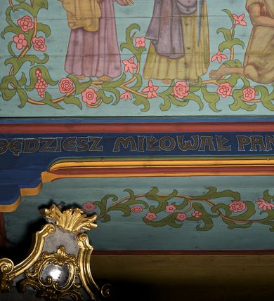 Zdjęcie nr 12: Ściany kościoła dekorują przedstawienia świętych: św. Stanisław w stroju pontyfikalnym z pastorałem, wskrzeszający Piotrowina. Poniżej na banderoli napis „BŁOGOSŁAWIENI KTÓRZY ŁAKNĄ / I PRAGNĄ SPRAWIEDLIWOŚCI”; św. Jadwiga w koronie i płaszczu podszytym gronostajem, trzymająca skrzyneczkę z klejnotami oraz krzyż z herbami Polski i Litwy. Na banderoli napis „BŁOGOSŁAWIENI / POKÓJ CZYNIĄCY”; św. Jan Kanty błogosławiący dziewczynę z dzbanem podpisany „BŁOGOSŁAWIENI / MIŁOSIERNI”; św. w habicie, z krzyżem w ręce i w asyście dwóch aniołów - jednego z palmą męczeństwa, drugiego z płonącą pochodnią, pod nimi napis „BŁOGOSŁAWIENI KTÓRZY / CIERPIĄ PRZEŚLADOWANIA DLA SPRAWIEDLIWOŚCI”; Chrystus z sercem gorejącym. Po jego prawej stronie klęczący mężczyzna, po lewej dwie owieczki nad strumieniem. Scena podpisana: „KTÓRZY PRACUJECIE I / OBCIĄŻENI JESTEŚCIE / A JA WAS OCHŁODZĘ”; archanioł Gabriel z lilią powyżej napisu  „ZDROWAŚ MARIO”; archanioł Michał z trybularzem, ponad napisem „KTÓŻ JAK BÓG!”; św. Franciszek na tle przyrody. Jest ubrany w habit franciszkański, na głowie ma kaptur. Przy prawej nodze świętego wilk, w jeziorze ryby, na niebie ptaki. Na banderoli napis „BŁOGOSŁAWIENI UBODZY DUCHEM / ALBOWIEM TAKOWYCH JEST / KRÓLESTWO NIEBIESKIE”; św. Izydor Oracz ukazany w towarzystwie anioła orającego na polu na tle dzwonnicy. Poniżej napis „BŁOGOSŁAWIENI CISI / ALBOWIEM ONI / POSIĄDĄ ZIEMIĘ”. Poniżej postaci świętych fryz z przedstawieniem 14 stacji drogi krzyżowej na błękitnym tle. Na stropie nawy i prezbiterium, na błękitnym tle, wśród kwiatowego ornamentu na środku ukazany Chrystus Ukrzyżowany ponad kielichem, gołębica Ducha Świętego i koronowana Maria. Po ich obu stronach stoją święci mężowie i niewiasty z atrybutami. W nawie w kolejności do środka: św. Wojciech (?), św. Jacek z figurą Matki Boskiej i monstrancją, klęczący św. Kazimierz w stroju książęcym z lilią, św. Agnieszka z barankiem i św. Karolina Kózkówna z różańcem, klęcząca św. Rozalia z różami, św. Klara z cyborium, św. Weronika z chustą z wizerunkiem Chrystusa, klęcząca św. Cecylia z lirą, św. Józef z lilią, św. Paweł z mieczem, klęczący św. Jan Chrzciciel z krzyżem. W prezbiterium przedstawiono 12 apostołów, niektórzy z atrybutmi, które ich jednoznacznie identyfilują, np. św. Andrzej z krzyżem w kształcie litery X, inni z księgami lub zwojami.  