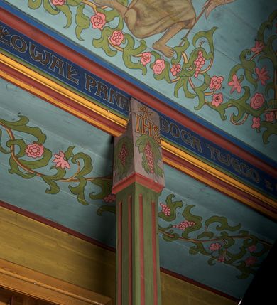 Zdjęcie nr 14: Ściany kościoła dekorują przedstawienia świętych: św. Stanisław w stroju pontyfikalnym z pastorałem, wskrzeszający Piotrowina. Poniżej na banderoli napis „BŁOGOSŁAWIENI KTÓRZY ŁAKNĄ / I PRAGNĄ SPRAWIEDLIWOŚCI”; św. Jadwiga w koronie i płaszczu podszytym gronostajem, trzymająca skrzyneczkę z klejnotami oraz krzyż z herbami Polski i Litwy. Na banderoli napis „BŁOGOSŁAWIENI / POKÓJ CZYNIĄCY”; św. Jan Kanty błogosławiący dziewczynę z dzbanem podpisany „BŁOGOSŁAWIENI / MIŁOSIERNI”; św. w habicie, z krzyżem w ręce i w asyście dwóch aniołów - jednego z palmą męczeństwa, drugiego z płonącą pochodnią, pod nimi napis „BŁOGOSŁAWIENI KTÓRZY / CIERPIĄ PRZEŚLADOWANIA DLA SPRAWIEDLIWOŚCI”; Chrystus z sercem gorejącym. Po jego prawej stronie klęczący mężczyzna, po lewej dwie owieczki nad strumieniem. Scena podpisana: „KTÓRZY PRACUJECIE I / OBCIĄŻENI JESTEŚCIE / A JA WAS OCHŁODZĘ”; archanioł Gabriel z lilią powyżej napisu  „ZDROWAŚ MARIO”; archanioł Michał z trybularzem, ponad napisem „KTÓŻ JAK BÓG!”; św. Franciszek na tle przyrody. Jest ubrany w habit franciszkański, na głowie ma kaptur. Przy prawej nodze świętego wilk, w jeziorze ryby, na niebie ptaki. Na banderoli napis „BŁOGOSŁAWIENI UBODZY DUCHEM / ALBOWIEM TAKOWYCH JEST / KRÓLESTWO NIEBIESKIE”; św. Izydor Oracz ukazany w towarzystwie anioła orającego na polu na tle dzwonnicy. Poniżej napis „BŁOGOSŁAWIENI CISI / ALBOWIEM ONI / POSIĄDĄ ZIEMIĘ”. Poniżej postaci świętych fryz z przedstawieniem 14 stacji drogi krzyżowej na błękitnym tle. Na stropie nawy i prezbiterium, na błękitnym tle, wśród kwiatowego ornamentu na środku ukazany Chrystus Ukrzyżowany ponad kielichem, gołębica Ducha Świętego i koronowana Maria. Po ich obu stronach stoją święci mężowie i niewiasty z atrybutami. W nawie w kolejności do środka: św. Wojciech (?), św. Jacek z figurą Matki Boskiej i monstrancją, klęczący św. Kazimierz w stroju książęcym z lilią, św. Agnieszka z barankiem i św. Karolina Kózkówna z różańcem, klęcząca św. Rozalia z różami, św. Klara z cyborium, św. Weronika z chustą z wizerunkiem Chrystusa, klęcząca św. Cecylia z lirą, św. Józef z lilią, św. Paweł z mieczem, klęczący św. Jan Chrzciciel z krzyżem. W prezbiterium przedstawiono 12 apostołów, niektórzy z atrybutmi, które ich jednoznacznie identyfilują, np. św. Andrzej z krzyżem w kształcie litery X, inni z księgami lub zwojami.  