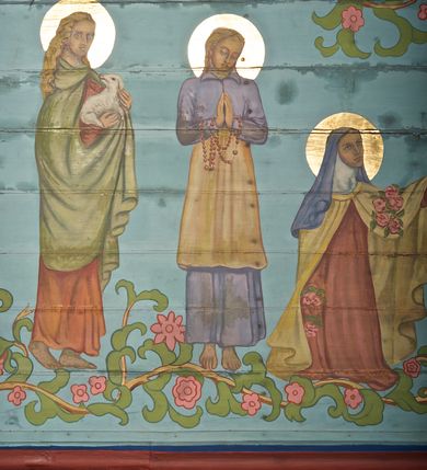 Zdjęcie nr 19: Ściany kościoła dekorują przedstawienia świętych: św. Stanisław w stroju pontyfikalnym z pastorałem, wskrzeszający Piotrowina. Poniżej na banderoli napis „BŁOGOSŁAWIENI KTÓRZY ŁAKNĄ / I PRAGNĄ SPRAWIEDLIWOŚCI”; św. Jadwiga w koronie i płaszczu podszytym gronostajem, trzymająca skrzyneczkę z klejnotami oraz krzyż z herbami Polski i Litwy. Na banderoli napis „BŁOGOSŁAWIENI / POKÓJ CZYNIĄCY”; św. Jan Kanty błogosławiący dziewczynę z dzbanem podpisany „BŁOGOSŁAWIENI / MIŁOSIERNI”; św. w habicie, z krzyżem w ręce i w asyście dwóch aniołów - jednego z palmą męczeństwa, drugiego z płonącą pochodnią, pod nimi napis „BŁOGOSŁAWIENI KTÓRZY / CIERPIĄ PRZEŚLADOWANIA DLA SPRAWIEDLIWOŚCI”; Chrystus z sercem gorejącym. Po jego prawej stronie klęczący mężczyzna, po lewej dwie owieczki nad strumieniem. Scena podpisana: „KTÓRZY PRACUJECIE I / OBCIĄŻENI JESTEŚCIE / A JA WAS OCHŁODZĘ”; archanioł Gabriel z lilią powyżej napisu  „ZDROWAŚ MARIO”; archanioł Michał z trybularzem, ponad napisem „KTÓŻ JAK BÓG!”; św. Franciszek na tle przyrody. Jest ubrany w habit franciszkański, na głowie ma kaptur. Przy prawej nodze świętego wilk, w jeziorze ryby, na niebie ptaki. Na banderoli napis „BŁOGOSŁAWIENI UBODZY DUCHEM / ALBOWIEM TAKOWYCH JEST / KRÓLESTWO NIEBIESKIE”; św. Izydor Oracz ukazany w towarzystwie anioła orającego na polu na tle dzwonnicy. Poniżej napis „BŁOGOSŁAWIENI CISI / ALBOWIEM ONI / POSIĄDĄ ZIEMIĘ”. Poniżej postaci świętych fryz z przedstawieniem 14 stacji drogi krzyżowej na błękitnym tle. Na stropie nawy i prezbiterium, na błękitnym tle, wśród kwiatowego ornamentu na środku ukazany Chrystus Ukrzyżowany ponad kielichem, gołębica Ducha Świętego i koronowana Maria. Po ich obu stronach stoją święci mężowie i niewiasty z atrybutami. W nawie w kolejności do środka: św. Wojciech (?), św. Jacek z figurą Matki Boskiej i monstrancją, klęczący św. Kazimierz w stroju książęcym z lilią, św. Agnieszka z barankiem i św. Karolina Kózkówna z różańcem, klęcząca św. Rozalia z różami, św. Klara z cyborium, św. Weronika z chustą z wizerunkiem Chrystusa, klęcząca św. Cecylia z lirą, św. Józef z lilią, św. Paweł z mieczem, klęczący św. Jan Chrzciciel z krzyżem. W prezbiterium przedstawiono 12 apostołów, niektórzy z atrybutmi, które ich jednoznacznie identyfilują, np. św. Andrzej z krzyżem w kształcie litery X, inni z księgami lub zwojami.  