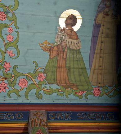 Zdjęcie nr 23: Ściany kościoła dekorują przedstawienia świętych: św. Stanisław w stroju pontyfikalnym z pastorałem, wskrzeszający Piotrowina. Poniżej na banderoli napis „BŁOGOSŁAWIENI KTÓRZY ŁAKNĄ / I PRAGNĄ SPRAWIEDLIWOŚCI”; św. Jadwiga w koronie i płaszczu podszytym gronostajem, trzymająca skrzyneczkę z klejnotami oraz krzyż z herbami Polski i Litwy. Na banderoli napis „BŁOGOSŁAWIENI / POKÓJ CZYNIĄCY”; św. Jan Kanty błogosławiący dziewczynę z dzbanem podpisany „BŁOGOSŁAWIENI / MIŁOSIERNI”; św. w habicie, z krzyżem w ręce i w asyście dwóch aniołów - jednego z palmą męczeństwa, drugiego z płonącą pochodnią, pod nimi napis „BŁOGOSŁAWIENI KTÓRZY / CIERPIĄ PRZEŚLADOWANIA DLA SPRAWIEDLIWOŚCI”; Chrystus z sercem gorejącym. Po jego prawej stronie klęczący mężczyzna, po lewej dwie owieczki nad strumieniem. Scena podpisana: „KTÓRZY PRACUJECIE I / OBCIĄŻENI JESTEŚCIE / A JA WAS OCHŁODZĘ”; archanioł Gabriel z lilią powyżej napisu  „ZDROWAŚ MARIO”; archanioł Michał z trybularzem, ponad napisem „KTÓŻ JAK BÓG!”; św. Franciszek na tle przyrody. Jest ubrany w habit franciszkański, na głowie ma kaptur. Przy prawej nodze świętego wilk, w jeziorze ryby, na niebie ptaki. Na banderoli napis „BŁOGOSŁAWIENI UBODZY DUCHEM / ALBOWIEM TAKOWYCH JEST / KRÓLESTWO NIEBIESKIE”; św. Izydor Oracz ukazany w towarzystwie anioła orającego na polu na tle dzwonnicy. Poniżej napis „BŁOGOSŁAWIENI CISI / ALBOWIEM ONI / POSIĄDĄ ZIEMIĘ”. Poniżej postaci świętych fryz z przedstawieniem 14 stacji drogi krzyżowej na błękitnym tle. Na stropie nawy i prezbiterium, na błękitnym tle, wśród kwiatowego ornamentu na środku ukazany Chrystus Ukrzyżowany ponad kielichem, gołębica Ducha Świętego i koronowana Maria. Po ich obu stronach stoją święci mężowie i niewiasty z atrybutami. W nawie w kolejności do środka: św. Wojciech (?), św. Jacek z figurą Matki Boskiej i monstrancją, klęczący św. Kazimierz w stroju książęcym z lilią, św. Agnieszka z barankiem i św. Karolina Kózkówna z różańcem, klęcząca św. Rozalia z różami, św. Klara z cyborium, św. Weronika z chustą z wizerunkiem Chrystusa, klęcząca św. Cecylia z lirą, św. Józef z lilią, św. Paweł z mieczem, klęczący św. Jan Chrzciciel z krzyżem. W prezbiterium przedstawiono 12 apostołów, niektórzy z atrybutmi, które ich jednoznacznie identyfilują, np. św. Andrzej z krzyżem w kształcie litery X, inni z księgami lub zwojami.  