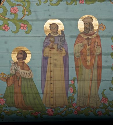 Zdjęcie nr 21: Ściany kościoła dekorują przedstawienia świętych: św. Stanisław w stroju pontyfikalnym z pastorałem, wskrzeszający Piotrowina. Poniżej na banderoli napis „BŁOGOSŁAWIENI KTÓRZY ŁAKNĄ / I PRAGNĄ SPRAWIEDLIWOŚCI”; św. Jadwiga w koronie i płaszczu podszytym gronostajem, trzymająca skrzyneczkę z klejnotami oraz krzyż z herbami Polski i Litwy. Na banderoli napis „BŁOGOSŁAWIENI / POKÓJ CZYNIĄCY”; św. Jan Kanty błogosławiący dziewczynę z dzbanem podpisany „BŁOGOSŁAWIENI / MIŁOSIERNI”; św. w habicie, z krzyżem w ręce i w asyście dwóch aniołów - jednego z palmą męczeństwa, drugiego z płonącą pochodnią, pod nimi napis „BŁOGOSŁAWIENI KTÓRZY / CIERPIĄ PRZEŚLADOWANIA DLA SPRAWIEDLIWOŚCI”; Chrystus z sercem gorejącym. Po jego prawej stronie klęczący mężczyzna, po lewej dwie owieczki nad strumieniem. Scena podpisana: „KTÓRZY PRACUJECIE I / OBCIĄŻENI JESTEŚCIE / A JA WAS OCHŁODZĘ”; archanioł Gabriel z lilią powyżej napisu  „ZDROWAŚ MARIO”; archanioł Michał z trybularzem, ponad napisem „KTÓŻ JAK BÓG!”; św. Franciszek na tle przyrody. Jest ubrany w habit franciszkański, na głowie ma kaptur. Przy prawej nodze świętego wilk, w jeziorze ryby, na niebie ptaki. Na banderoli napis „BŁOGOSŁAWIENI UBODZY DUCHEM / ALBOWIEM TAKOWYCH JEST / KRÓLESTWO NIEBIESKIE”; św. Izydor Oracz ukazany w towarzystwie anioła orającego na polu na tle dzwonnicy. Poniżej napis „BŁOGOSŁAWIENI CISI / ALBOWIEM ONI / POSIĄDĄ ZIEMIĘ”. Poniżej postaci świętych fryz z przedstawieniem 14 stacji drogi krzyżowej na błękitnym tle. Na stropie nawy i prezbiterium, na błękitnym tle, wśród kwiatowego ornamentu na środku ukazany Chrystus Ukrzyżowany ponad kielichem, gołębica Ducha Świętego i koronowana Maria. Po ich obu stronach stoją święci mężowie i niewiasty z atrybutami. W nawie w kolejności do środka: św. Wojciech (?), św. Jacek z figurą Matki Boskiej i monstrancją, klęczący św. Kazimierz w stroju książęcym z lilią, św. Agnieszka z barankiem i św. Karolina Kózkówna z różańcem, klęcząca św. Rozalia z różami, św. Klara z cyborium, św. Weronika z chustą z wizerunkiem Chrystusa, klęcząca św. Cecylia z lirą, św. Józef z lilią, św. Paweł z mieczem, klęczący św. Jan Chrzciciel z krzyżem. W prezbiterium przedstawiono 12 apostołów, niektórzy z atrybutmi, które ich jednoznacznie identyfilują, np. św. Andrzej z krzyżem w kształcie litery X, inni z księgami lub zwojami.  