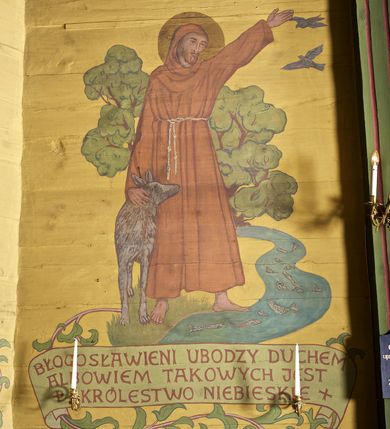 Zdjęcie nr 5: Ściany kościoła dekorują przedstawienia świętych: św. Stanisław w stroju pontyfikalnym z pastorałem, wskrzeszający Piotrowina. Poniżej na banderoli napis „BŁOGOSŁAWIENI KTÓRZY ŁAKNĄ / I PRAGNĄ SPRAWIEDLIWOŚCI”; św. Jadwiga w koronie i płaszczu podszytym gronostajem, trzymająca skrzyneczkę z klejnotami oraz krzyż z herbami Polski i Litwy. Na banderoli napis „BŁOGOSŁAWIENI / POKÓJ CZYNIĄCY”; św. Jan Kanty błogosławiący dziewczynę z dzbanem podpisany „BŁOGOSŁAWIENI / MIŁOSIERNI”; św. w habicie, z krzyżem w ręce i w asyście dwóch aniołów - jednego z palmą męczeństwa, drugiego z płonącą pochodnią, pod nimi napis „BŁOGOSŁAWIENI KTÓRZY / CIERPIĄ PRZEŚLADOWANIA DLA SPRAWIEDLIWOŚCI”; Chrystus z sercem gorejącym. Po jego prawej stronie klęczący mężczyzna, po lewej dwie owieczki nad strumieniem. Scena podpisana: „KTÓRZY PRACUJECIE I / OBCIĄŻENI JESTEŚCIE / A JA WAS OCHŁODZĘ”; archanioł Gabriel z lilią powyżej napisu  „ZDROWAŚ MARIO”; archanioł Michał z trybularzem, ponad napisem „KTÓŻ JAK BÓG!”; św. Franciszek na tle przyrody. Jest ubrany w habit franciszkański, na głowie ma kaptur. Przy prawej nodze świętego wilk, w jeziorze ryby, na niebie ptaki. Na banderoli napis „BŁOGOSŁAWIENI UBODZY DUCHEM / ALBOWIEM TAKOWYCH JEST / KRÓLESTWO NIEBIESKIE”; św. Izydor Oracz ukazany w towarzystwie anioła orającego na polu na tle dzwonnicy. Poniżej napis „BŁOGOSŁAWIENI CISI / ALBOWIEM ONI / POSIĄDĄ ZIEMIĘ”. Poniżej postaci świętych fryz z przedstawieniem 14 stacji drogi krzyżowej na błękitnym tle. Na stropie nawy i prezbiterium, na błękitnym tle, wśród kwiatowego ornamentu na środku ukazany Chrystus Ukrzyżowany ponad kielichem, gołębica Ducha Świętego i koronowana Maria. Po ich obu stronach stoją święci mężowie i niewiasty z atrybutami. W nawie w kolejności do środka: św. Wojciech (?), św. Jacek z figurą Matki Boskiej i monstrancją, klęczący św. Kazimierz w stroju książęcym z lilią, św. Agnieszka z barankiem i św. Karolina Kózkówna z różańcem, klęcząca św. Rozalia z różami, św. Klara z cyborium, św. Weronika z chustą z wizerunkiem Chrystusa, klęcząca św. Cecylia z lirą, św. Józef z lilią, św. Paweł z mieczem, klęczący św. Jan Chrzciciel z krzyżem. W prezbiterium przedstawiono 12 apostołów, niektórzy z atrybutmi, które ich jednoznacznie identyfilują, np. św. Andrzej z krzyżem w kształcie litery X, inni z księgami lub zwojami.  