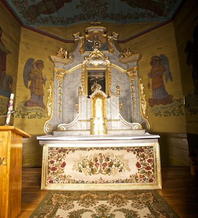 Zdjęcie nr 1: Ołtarz architektoniczny, jednoosiowy, jednokondygnacyjny. Mensa prostopadłościenna z antepedium haftowanym bogatą dekoracją kwiatową, z centralnie umieszczonym kielichem w otoczeniu winnej latorośli. Na nim tabernakulum w formie zbliżonej do prostopadłościennej skrzynki o wypukłej ściance frontowej na tle ścianki z tronem wystawienia ustawionej na cokole i ujętej spływami wolutowymi. Drzwiczki tabernakulum dekorowane krzyżem z glorią między literami „Α” i „Ω”. Tron wystawienia złocony, dekorowany kwiatem między stylizowanymi liśćmi i ujęty od góry muszlą palmetową. Tron ujmują lizeny przedzielone i zwieńczone gzymsem ustawionym skośnie i zakończonym od wewnętrznej strony wolutami. Gzyms nad tronem wystawienia wyniesiony, w formie łuku nadwieszonego. Na jego zewnętrznych częściach posadowiono figurki klęczących i modlących się aniołków. Nastawa ujęta parą zdwojonych pilastrów na cokołach, o głowicach korynckich, z impostami z dekoracją w formie festonów z rocaille’em i liśćmi. Uszy w formie spływów wolutowych z dekoracją liściastą. W centrum pole obrazowe z wnęką o profilowanej ramie w kształcie stojącego prostokąta ujętego w spływy wolutowe i festony, zwieńczone wydatnym kartuszem dekorowanym rocaille’em, z monogramem maryjnym. W nim ukazana Matka Boska Anielska przesłaniana obrazem „Wszyscy Święci” wzorowanym na obrazie Rafaela. Retabulum przedzielone gzymsem wyniesionym w łuk ponad polem centralnym. Zwieńczenie w formie zbliżonej do prostokąta, rozszerzające się ku dołowi, ujęte w esowate pilastry przechodzące w woluty, zamknięte łukiem odcinkowym zakończonym wolutami. Całość wieńczy krzyż z rozetą na skrzyżowaniu ramion. W zwieńczeniu wklęsło-wypukłe pole obrazowe z płaskorzeźbionym popiersiem Boga Ojca na tle nieba. Na impostach leżące figury aniołów. Struktura polichromowana, marmoryzowana w odcieniu jasnoszarym, profile, ornamenty i dekoracje złocone i srebrzone. 