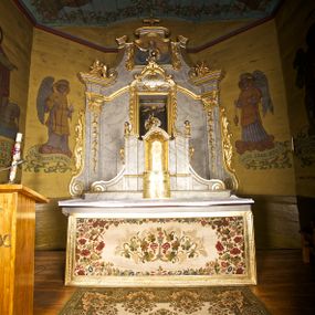 Zdjęcie nr 1: Ołtarz architektoniczny, jednoosiowy, jednokondygnacyjny. Mensa prostopadłościenna z antepedium haftowanym bogatą dekoracją kwiatową, z centralnie umieszczonym kielichem w otoczeniu winnej latorośli. Na nim tabernakulum w formie zbliżonej do prostopadłościennej skrzynki o wypukłej ściance frontowej na tle ścianki z tronem wystawienia ustawionej na cokole i ujętej spływami wolutowymi. Drzwiczki tabernakulum dekorowane krzyżem z glorią między literami „Α” i „Ω”. Tron wystawienia złocony, dekorowany kwiatem między stylizowanymi liśćmi i ujęty od góry muszlą palmetową. Tron ujmują lizeny przedzielone i zwieńczone gzymsem ustawionym skośnie i zakończonym od wewnętrznej strony wolutami. Gzyms nad tronem wystawienia wyniesiony, w formie łuku nadwieszonego. Na jego zewnętrznych częściach posadowiono figurki klęczących i modlących się aniołków. Nastawa ujęta parą zdwojonych pilastrów na cokołach, o głowicach korynckich, z impostami z dekoracją w formie festonów z rocaille’em i liśćmi. Uszy w formie spływów wolutowych z dekoracją liściastą. W centrum pole obrazowe z wnęką o profilowanej ramie w kształcie stojącego prostokąta ujętego w spływy wolutowe i festony, zwieńczone wydatnym kartuszem dekorowanym rocaille’em, z monogramem maryjnym. W nim ukazana Matka Boska Anielska przesłaniana obrazem „Wszyscy Święci” wzorowanym na obrazie Rafaela. Retabulum przedzielone gzymsem wyniesionym w łuk ponad polem centralnym. Zwieńczenie w formie zbliżonej do prostokąta, rozszerzające się ku dołowi, ujęte w esowate pilastry przechodzące w woluty, zamknięte łukiem odcinkowym zakończonym wolutami. Całość wieńczy krzyż z rozetą na skrzyżowaniu ramion. W zwieńczeniu wklęsło-wypukłe pole obrazowe z płaskorzeźbionym popiersiem Boga Ojca na tle nieba. Na impostach leżące figury aniołów. Struktura polichromowana, marmoryzowana w odcieniu jasnoszarym, profile, ornamenty i dekoracje złocone i srebrzone. 