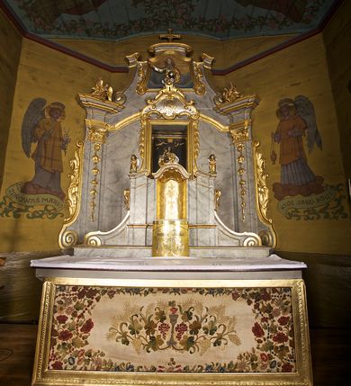 Zdjęcie nr 2: Tabernakulum w formie zbliżonej do prostopadłościennej skrzynki o wypukłej ściance frontowej ustawione na tle ścianki z tronem wystawienia ustawionej na cokole i ujętej spływami wolutowymi.  Drzwiczki tabernakulum dekorowane krzyżem z glorią między literami „Α” i „Ω”. Tron wystawienia złocony, dekorowany kwiatem między stylizowanymi liśćmi i zamknięty od góry muszlą palmetową. Tron ujmują lizeny przedzielone i zwieńczone gzymsem ustawionym skośnie i zakończonym od wewnętrznej strony wolutami. Gzyms nad tronem wystawienia wyniesiony, w formie łuku nadwieszonego. Na jego zewnętrznych częściach posadowiono figurki klęczących i modlących się aniołków. Tabernakulum polichromowane na kolor jasnoszary, marmoryzowane, profile złocone. 