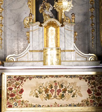 Zdjęcie nr 1: Tabernakulum w formie zbliżonej do prostopadłościennej skrzynki o wypukłej ściance frontowej ustawione na tle ścianki z tronem wystawienia ustawionej na cokole i ujętej spływami wolutowymi.  Drzwiczki tabernakulum dekorowane krzyżem z glorią między literami „Α” i „Ω”. Tron wystawienia złocony, dekorowany kwiatem między stylizowanymi liśćmi i zamknięty od góry muszlą palmetową. Tron ujmują lizeny przedzielone i zwieńczone gzymsem ustawionym skośnie i zakończonym od wewnętrznej strony wolutami. Gzyms nad tronem wystawienia wyniesiony, w formie łuku nadwieszonego. Na jego zewnętrznych częściach posadowiono figurki klęczących i modlących się aniołków. Tabernakulum polichromowane na kolor jasnoszary, marmoryzowane, profile złocone. 