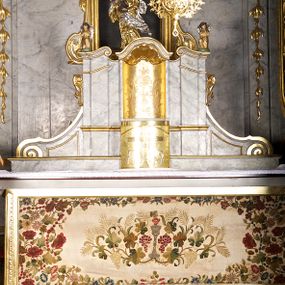 Zdjęcie nr 1: Tabernakulum w formie zbliżonej do prostopadłościennej skrzynki o wypukłej ściance frontowej ustawione na tle ścianki z tronem wystawienia ustawionej na cokole i ujętej spływami wolutowymi.  Drzwiczki tabernakulum dekorowane krzyżem z glorią między literami „Α” i „Ω”. Tron wystawienia złocony, dekorowany kwiatem między stylizowanymi liśćmi i zamknięty od góry muszlą palmetową. Tron ujmują lizeny przedzielone i zwieńczone gzymsem ustawionym skośnie i zakończonym od wewnętrznej strony wolutami. Gzyms nad tronem wystawienia wyniesiony, w formie łuku nadwieszonego. Na jego zewnętrznych częściach posadowiono figurki klęczących i modlących się aniołków. Tabernakulum polichromowane na kolor jasnoszary, marmoryzowane, profile złocone. 