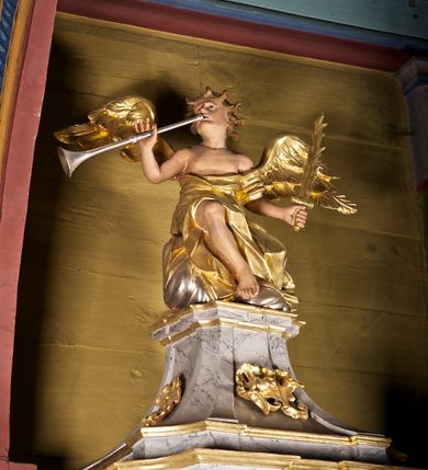 Zdjęcie nr 4: Ambona przyścienna składająca się z kosza, zaplecka i baldachimu zawieszona na lewej ścianie prezbiterium. Kosz na planie kwadratu, o wklęsłych narożach dekorowanych rocaille'em i ujętych pilastrami, dekorowany ramami o krótkich krawędziach utworzonych z ornamentu rocaille’owego; parapet profilowany, wklęsło-wypukły. Podstawę tworzy stylizowany motyw muszli, z podwieszonym ośmiobokiem zakończonym kwiatonem. Zaplecek gładki, zamknięty belkowaniem, podtrzymany na konsolach, z których zwiesza się ornament roślinny. W dolnej części płycina w kształcie leżącego prostokąta. Baldachim powtarzający kształt kosza, w formie bogato profilowanego gzymsu z gołębicą w glorii na podniebieniu. Powyżej gzymsu baldachim przechodzi w zwężający się, dekolorowany rocaille'em cokół, na którym znajduje się pełnoplastyczna rzeźba anioła siedzącego na chmurze, dmącego w trąbę trzymaną w prawej ręce, lewą ujmującego płomienisty miecz. Anioł ukazany został w skręcie w prawo, z głową lekko uniesioną i rozłożonymi skrzydłami. Jest ubrany w obszerny, złoty płaszcz, spod którego wystaje prawa noga. Struktura marmoryzowana na kolor jasnoszary; ornamenty, profile i fragmenty rzeźb złocone, gołębica, chmura i trąba srebrzone. 