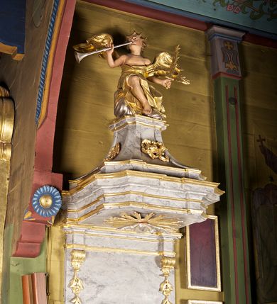 Zdjęcie nr 3: Ambona przyścienna składająca się z kosza, zaplecka i baldachimu zawieszona na lewej ścianie prezbiterium. Kosz na planie kwadratu, o wklęsłych narożach dekorowanych rocaille'em i ujętych pilastrami, dekorowany ramami o krótkich krawędziach utworzonych z ornamentu rocaille’owego; parapet profilowany, wklęsło-wypukły. Podstawę tworzy stylizowany motyw muszli, z podwieszonym ośmiobokiem zakończonym kwiatonem. Zaplecek gładki, zamknięty belkowaniem, podtrzymany na konsolach, z których zwiesza się ornament roślinny. W dolnej części płycina w kształcie leżącego prostokąta. Baldachim powtarzający kształt kosza, w formie bogato profilowanego gzymsu z gołębicą w glorii na podniebieniu. Powyżej gzymsu baldachim przechodzi w zwężający się, dekolorowany rocaille'em cokół, na którym znajduje się pełnoplastyczna rzeźba anioła siedzącego na chmurze, dmącego w trąbę trzymaną w prawej ręce, lewą ujmującego płomienisty miecz. Anioł ukazany został w skręcie w prawo, z głową lekko uniesioną i rozłożonymi skrzydłami. Jest ubrany w obszerny, złoty płaszcz, spod którego wystaje prawa noga. Struktura marmoryzowana na kolor jasnoszary; ornamenty, profile i fragmenty rzeźb złocone, gołębica, chmura i trąba srebrzone. 