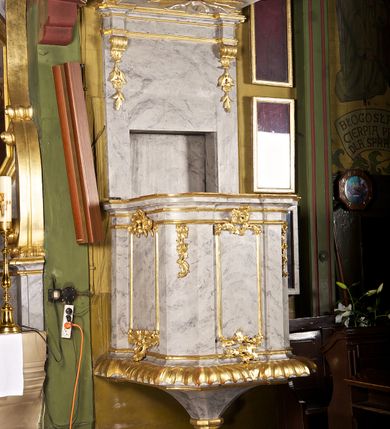 Zdjęcie nr 1: Ambona przyścienna składająca się z kosza, zaplecka i baldachimu zawieszona na lewej ścianie prezbiterium. Kosz na planie kwadratu, o wklęsłych narożach dekorowanych rocaille'em i ujętych pilastrami, dekorowany ramami o krótkich krawędziach utworzonych z ornamentu rocaille’owego; parapet profilowany, wklęsło-wypukły. Podstawę tworzy stylizowany motyw muszli, z podwieszonym ośmiobokiem zakończonym kwiatonem. Zaplecek gładki, zamknięty belkowaniem, podtrzymany na konsolach, z których zwiesza się ornament roślinny. W dolnej części płycina w kształcie leżącego prostokąta. Baldachim powtarzający kształt kosza, w formie bogato profilowanego gzymsu z gołębicą w glorii na podniebieniu. Powyżej gzymsu baldachim przechodzi w zwężający się, dekolorowany rocaille'em cokół, na którym znajduje się pełnoplastyczna rzeźba anioła siedzącego na chmurze, dmącego w trąbę trzymaną w prawej ręce, lewą ujmującego płomienisty miecz. Anioł ukazany został w skręcie w prawo, z głową lekko uniesioną i rozłożonymi skrzydłami. Jest ubrany w obszerny, złoty płaszcz, spod którego wystaje prawa noga. Struktura marmoryzowana na kolor jasnoszary; ornamenty, profile i fragmenty rzeźb złocone, gołębica, chmura i trąba srebrzone. 