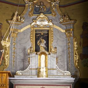 Zdjęcie nr 1: Obraz w kształcie stojącego prostokąta z przedstawieniem Matki Boskiej z Dzieciątkiem. W centrum kompozycji Matka Boska ujęta w całej postaci, frontalnie, podtrzymująca obiema rękami Dzieciątko. Maria na owalną twarz, wzrok kieruje w górę, ma wąski nos i pełne usta, długie włosy zaczesane do tyłu. Partie ubrań przykrywa rzeźbiona sukienka, złocona w obrębie sukni i srebrzona w części płaszcza. Na obu ornament floralny. Dzieciątko siedzi w ramionach Marii, jest pulchne, rączki rozkłada do boków, spogląda lekko w górę, na głowie ma zamkniętą, złotą koronę. Ponad głową Marii unoszą się symetrycznie dwa putta, z których każde w jednej ręce trzyma gałązkę kwitnącego kwiatu a drugą podtrzymuje koronę nad głową Niewiasty. Tak, jak w przypadku Dzieciątka, jest to korona zamknięta nałożona na obraz i wykonana z trybowanej, złoconej blachy. Tło obrazu jednolite, ciemne.   