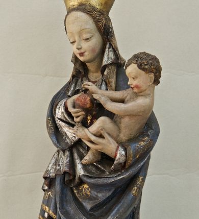 Zdjęcie nr 1: Rzeźba pełna, ukazująca Matkę Boską z Dzieciątkiem na niskiej, wielobocznej podstawie. Maria stoi, lewą ręką podtrzymuje Jezusa, a prawą podaje mu jabłko i spogląda na ten gest. Ma owalną, pełną twarz, o wysokim czole, wąskich brwiach, dużych, półprzymkniętych oczach, niewielkim nosie i ustach rozchylonych w delikatnym uśmiechu. Jest ubrana w czerwoną suknię ze złotym ornamentem, której fragment widać przy prawej nodze i bardzo obszerny, gęsto drapowany granatowy płaszcz ze złotym ornamentem i srebrnym podbiciem. Wierzchnie okrycie owija się wokół lewej ręki Madonny i szerokimi, twardo giętymi fałdami spływa w dół; przy prawym biodrze tworzy rurkowato zwinięte kaskady. Maria ma na głowę zarzucony welon, który, wywijając się, spływa na dekolt oraz wysoką, złotą, otwarta koronę. Rzeźba jest sygnowana z tyłu przy dolnej krawędzi „A(NDRZEJ) WALIJEWSKI”. Polichromia w partiach ciała naturalistyczna. 