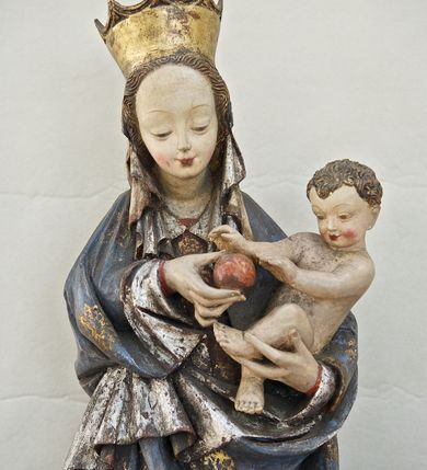 Zdjęcie nr 9: Rzeźba pełna, ukazująca Matkę Boską z Dzieciątkiem na niskiej, wielobocznej podstawie. Maria stoi, lewą ręką podtrzymuje Jezusa, a prawą podaje mu jabłko i spogląda na ten gest. Ma owalną, pełną twarz, o wysokim czole, wąskich brwiach, dużych, półprzymkniętych oczach, niewielkim nosie i ustach rozchylonych w delikatnym uśmiechu. Jest ubrana w czerwoną suknię ze złotym ornamentem, której fragment widać przy prawej nodze i bardzo obszerny, gęsto drapowany granatowy płaszcz ze złotym ornamentem i srebrnym podbiciem. Wierzchnie okrycie owija się wokół lewej ręki Madonny i szerokimi, twardo giętymi fałdami spływa w dół; przy prawym biodrze tworzy rurkowato zwinięte kaskady. Maria ma na głowę zarzucony welon, który, wywijając się, spływa na dekolt oraz wysoką, złotą, otwarta koronę. Rzeźba jest sygnowana z tyłu przy dolnej krawędzi „A(NDRZEJ) WALIJEWSKI”. Polichromia w partiach ciała naturalistyczna. 